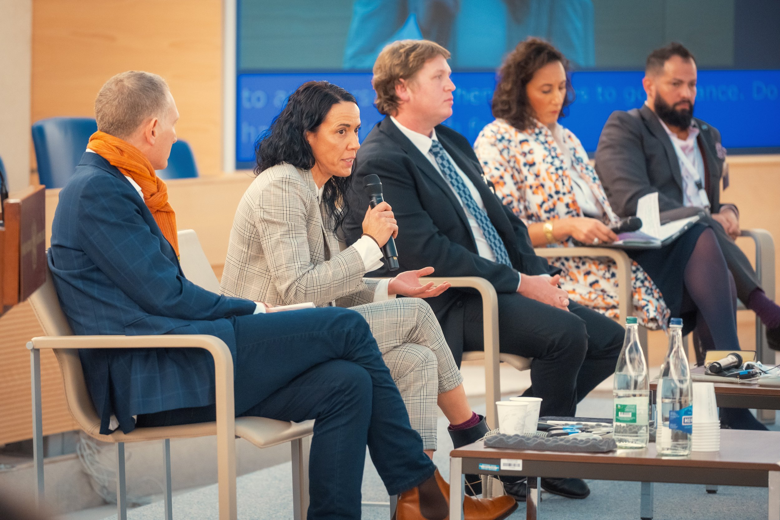 Panel of five diverse people seated at a conference table with water bottles, listening to a presentation in a modern conference room.