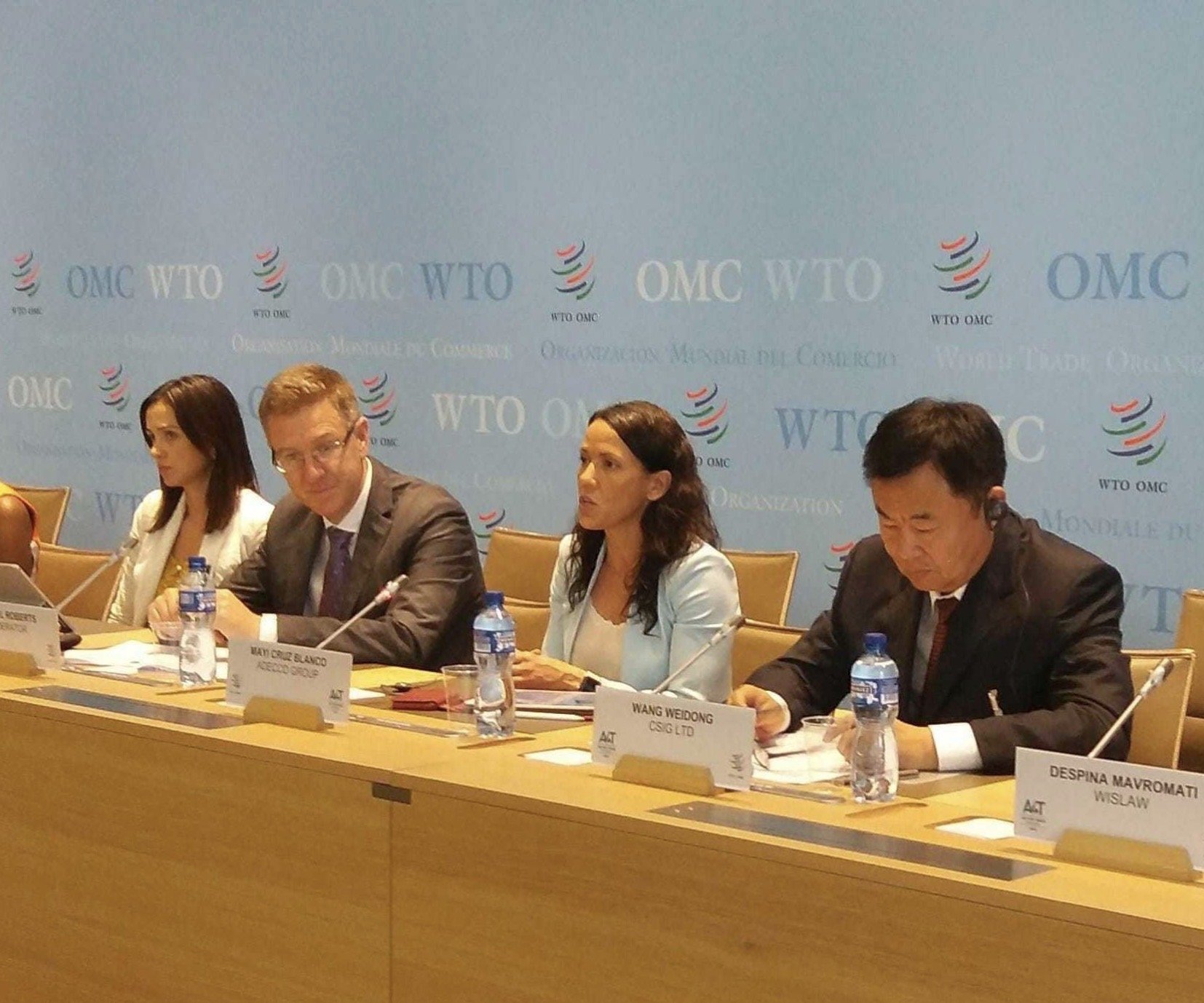 Four individuals sitting at a conference table with name tags and water bottles in front of them. Behind them is a blue banner with the WTO and WHO logos, and text that reads "OMC" and "Organisation Mondiale de Coopération".