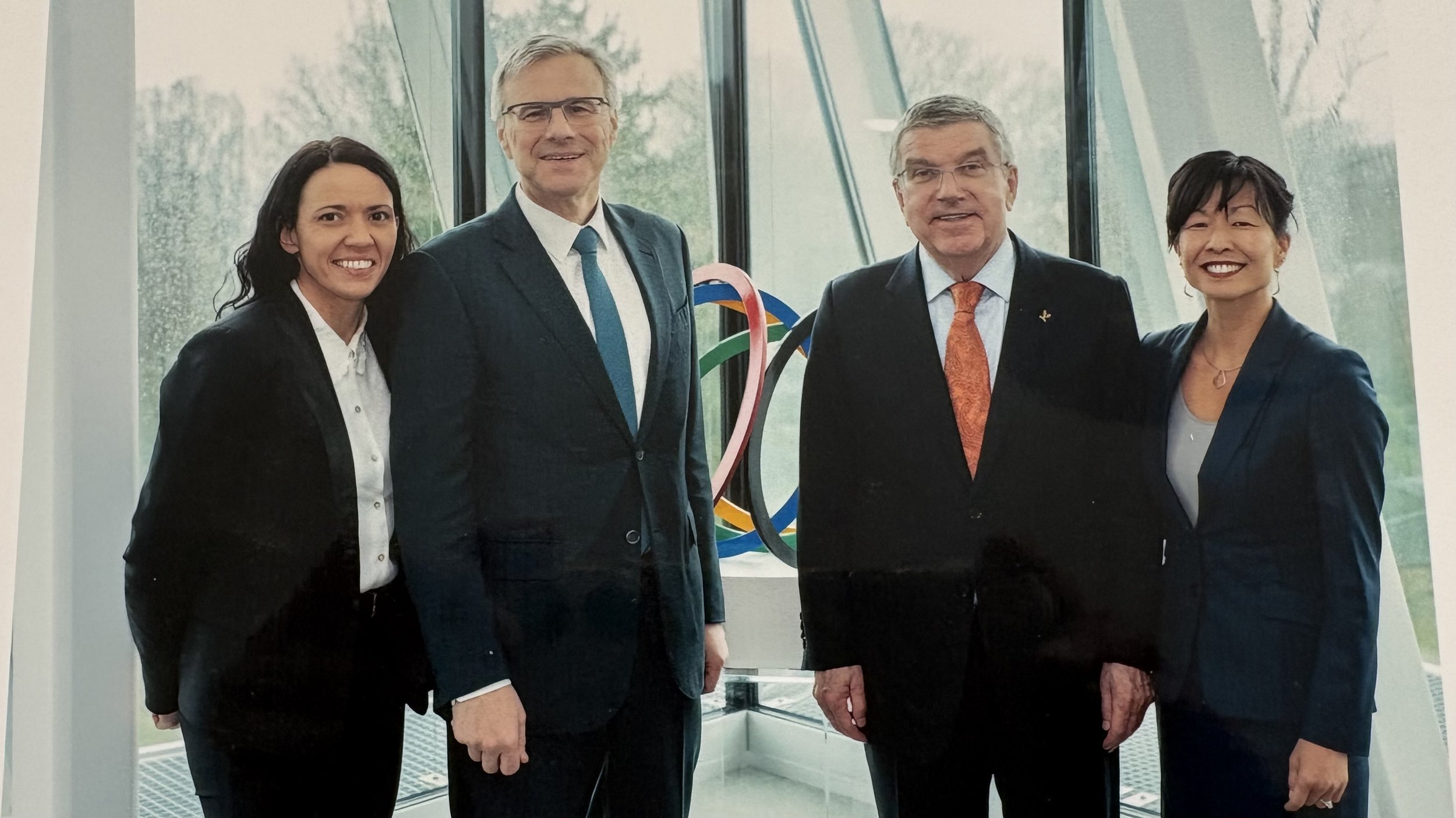 Four professionally dressed individuals standing together indoors with large window in the background and Olympic rings in the distance. From left to right: woman with dark hair, man with glasses and gray hair, man with gray hair and orange tie, woman with short dark hair.