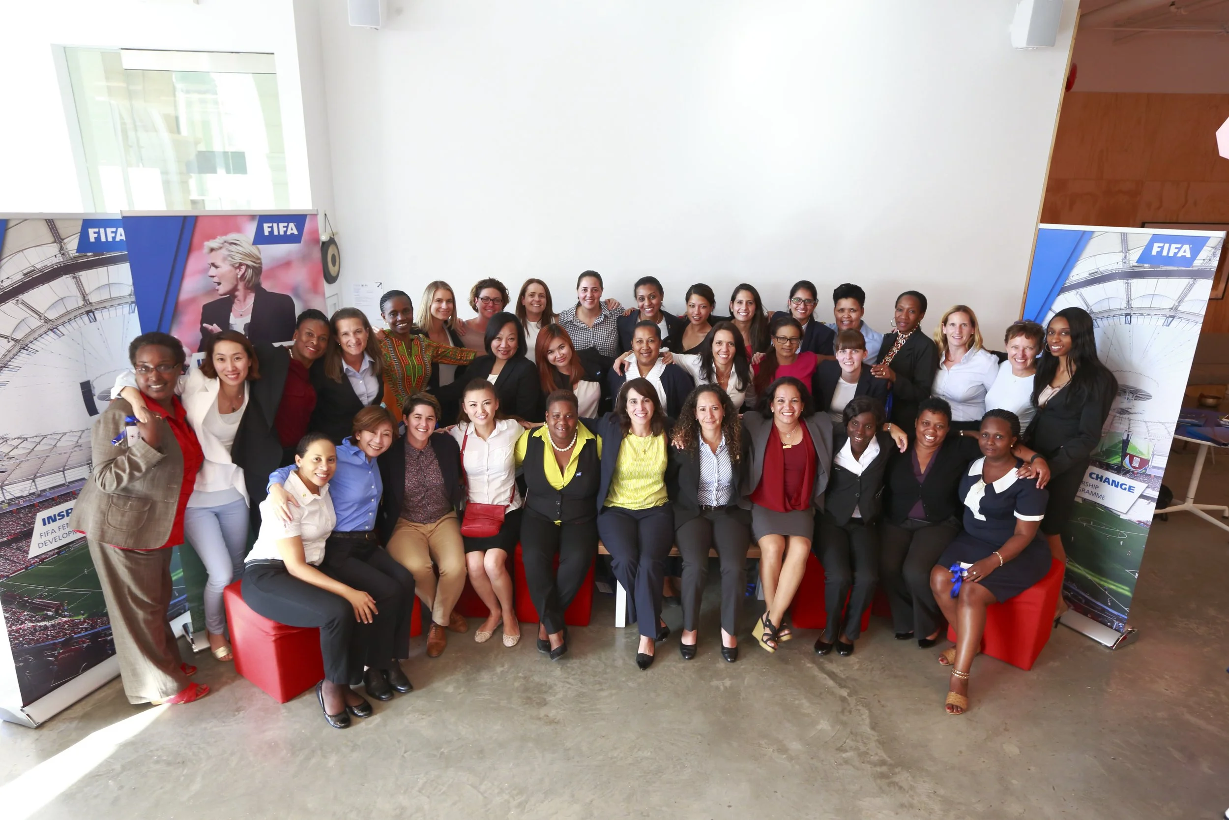 Group of women and men posing together indoors at an event, some standing and some sitting, with FIFA banners in the background.