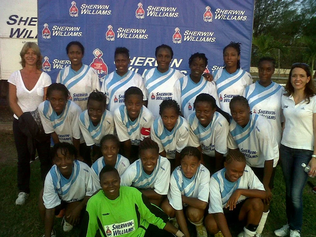 A group of young women soccer players in white and blue uniforms along with two adult women standing on either side, posing for a team photo outdoors in front of a blue Sherwin Williams backdrop.