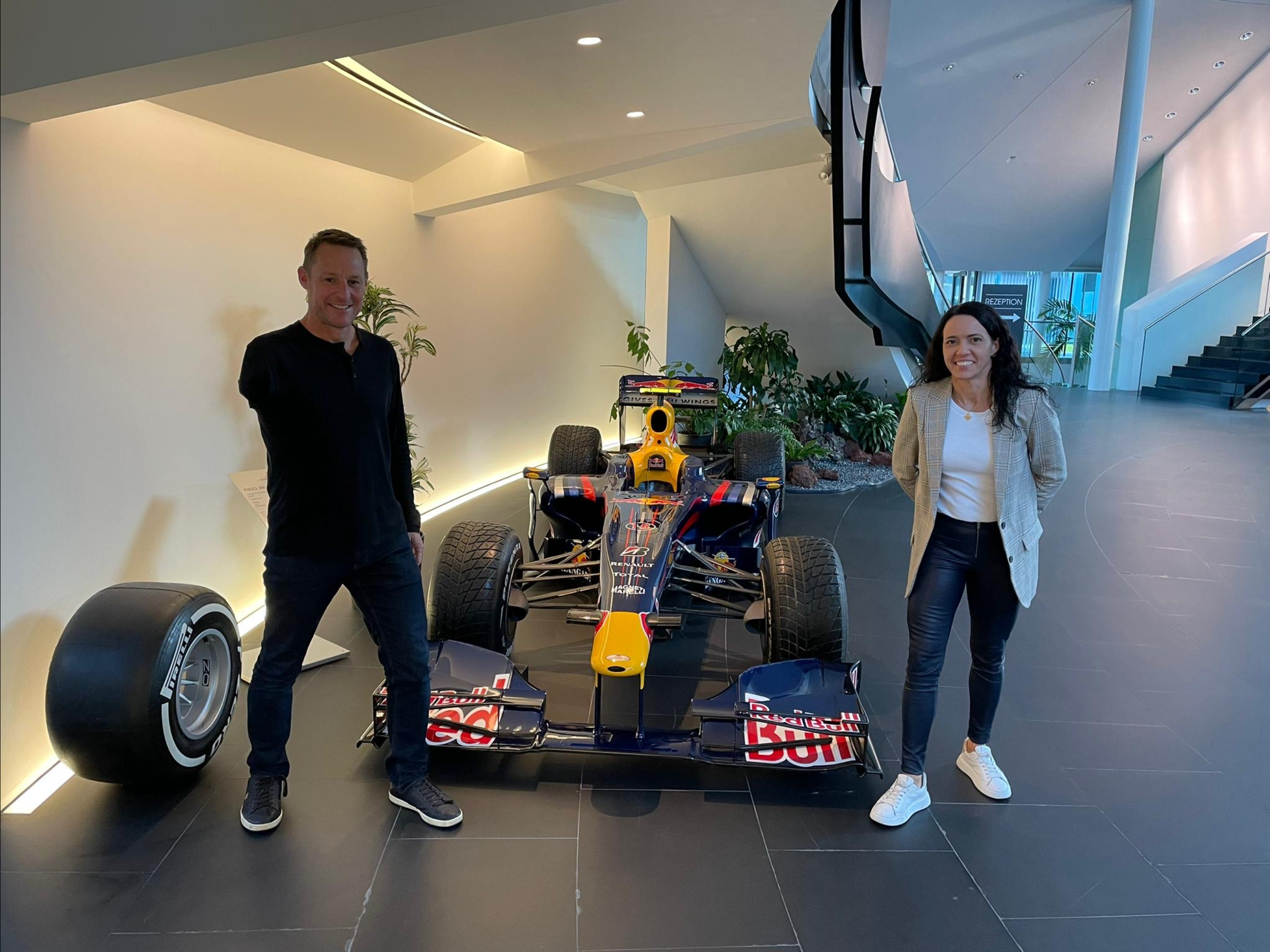 Two people standing next to a Red Bull Racing Formula 1 car in a modern indoor space with stairs and plants.