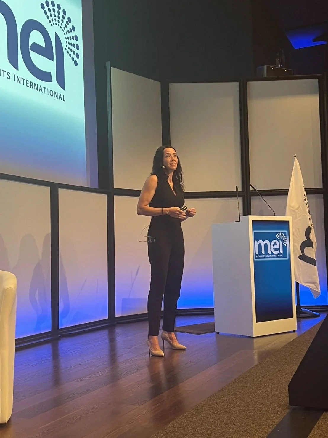 A woman with long dark hair wearing a black sleeveless top, black pants, and beige high heels speaking at a podium with a microphone. Behind her is a large screen displaying the logo of Major Events International (MEI).