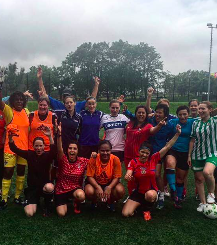 A group of young women and girls in sports uniforms posing together on a soccer field, celebrating with raised arms and smiles, after a game.
