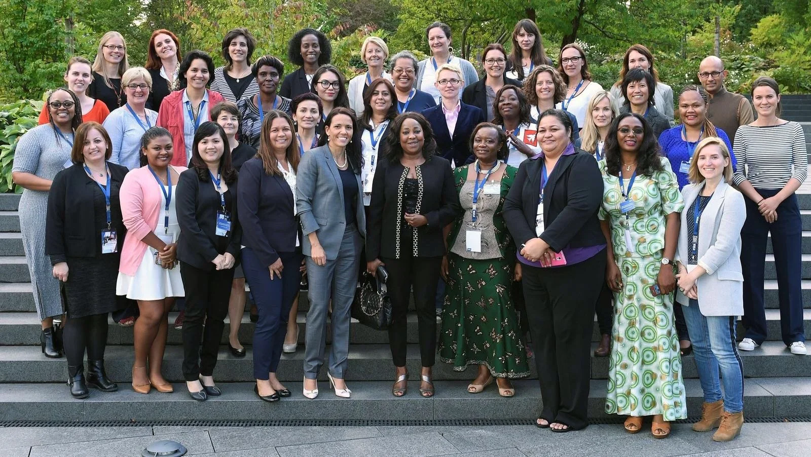 Group photo of diverse women and a few men standing on outdoor steps, smiling, with green trees in the background.