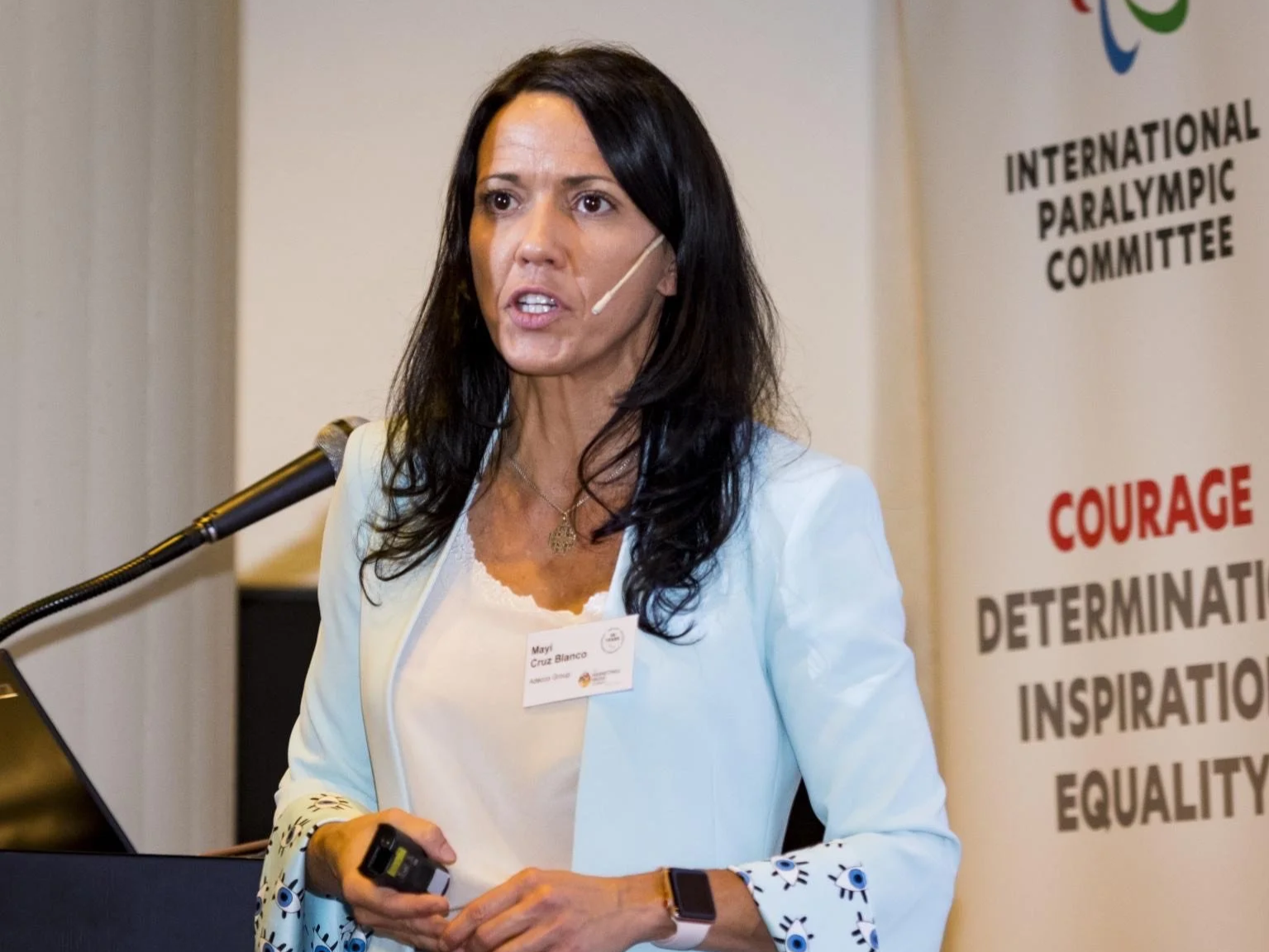 Woman giving a presentation at an event, wearing a white blazer and a name badge that reads 'May Cruz Blanco', with a banner in the background that reads 'International Paralympic Committee' and words like 'Courage', 'Determination', 'Inspiration', and 'Equality'.