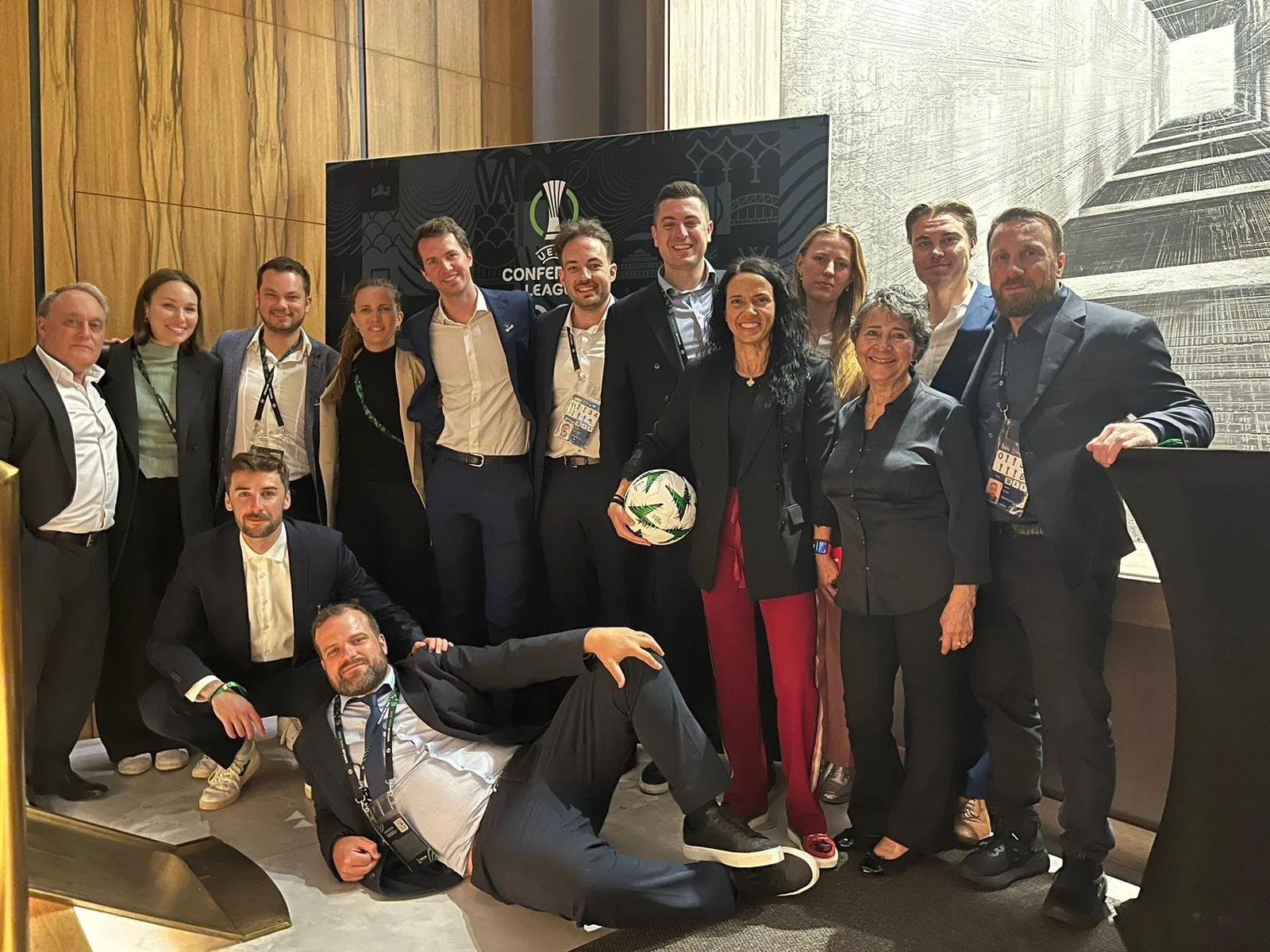 Group of fourteen people, mostly professionals in business attire, posing for a photo at a conference, with a logo and the words "Conference League" in the background.