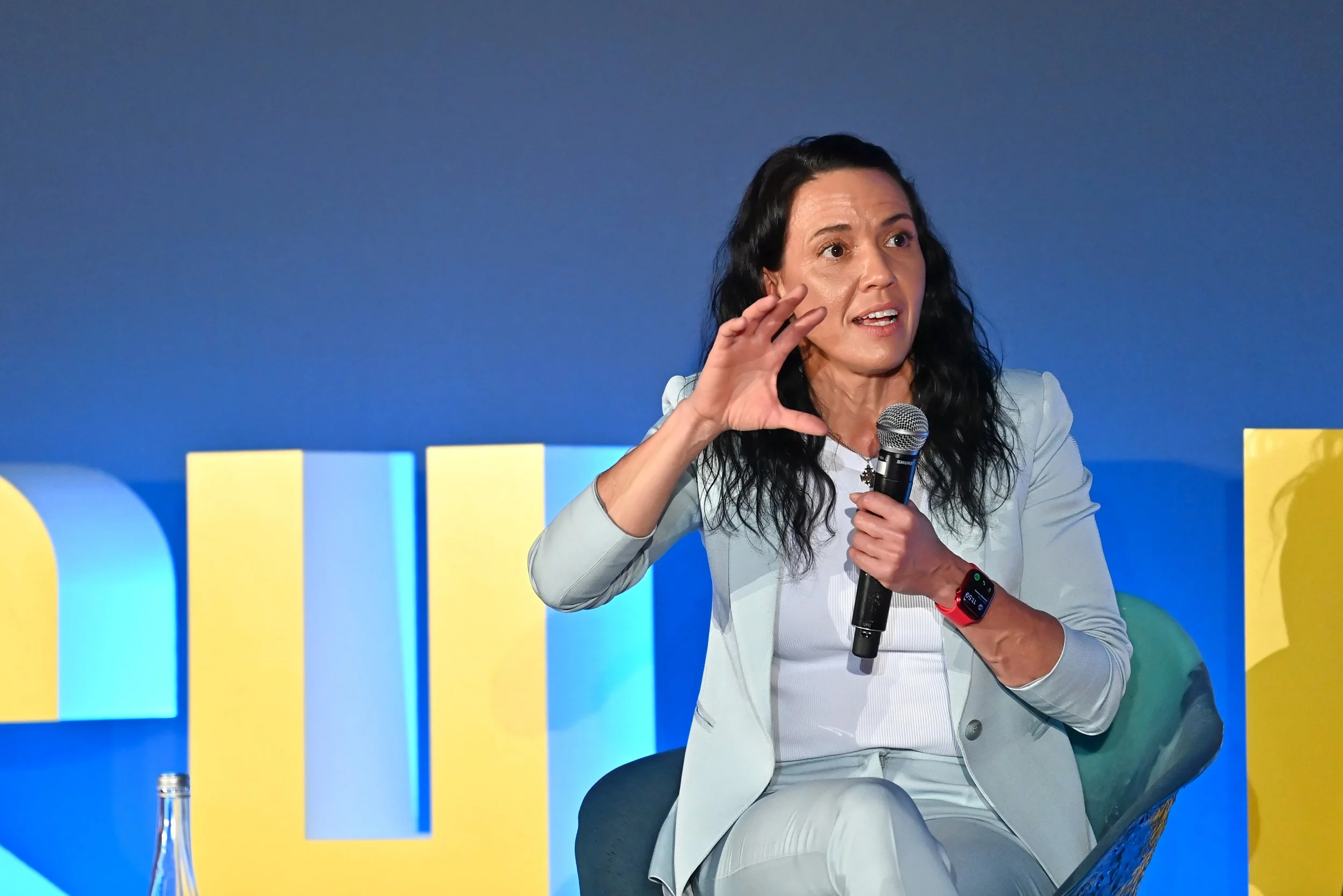 A woman in a light gray suit speaking into a microphone during a panel discussion or presentation. She has long dark hair and is gesturing with her right hand. The background features colorful geometric shapes.