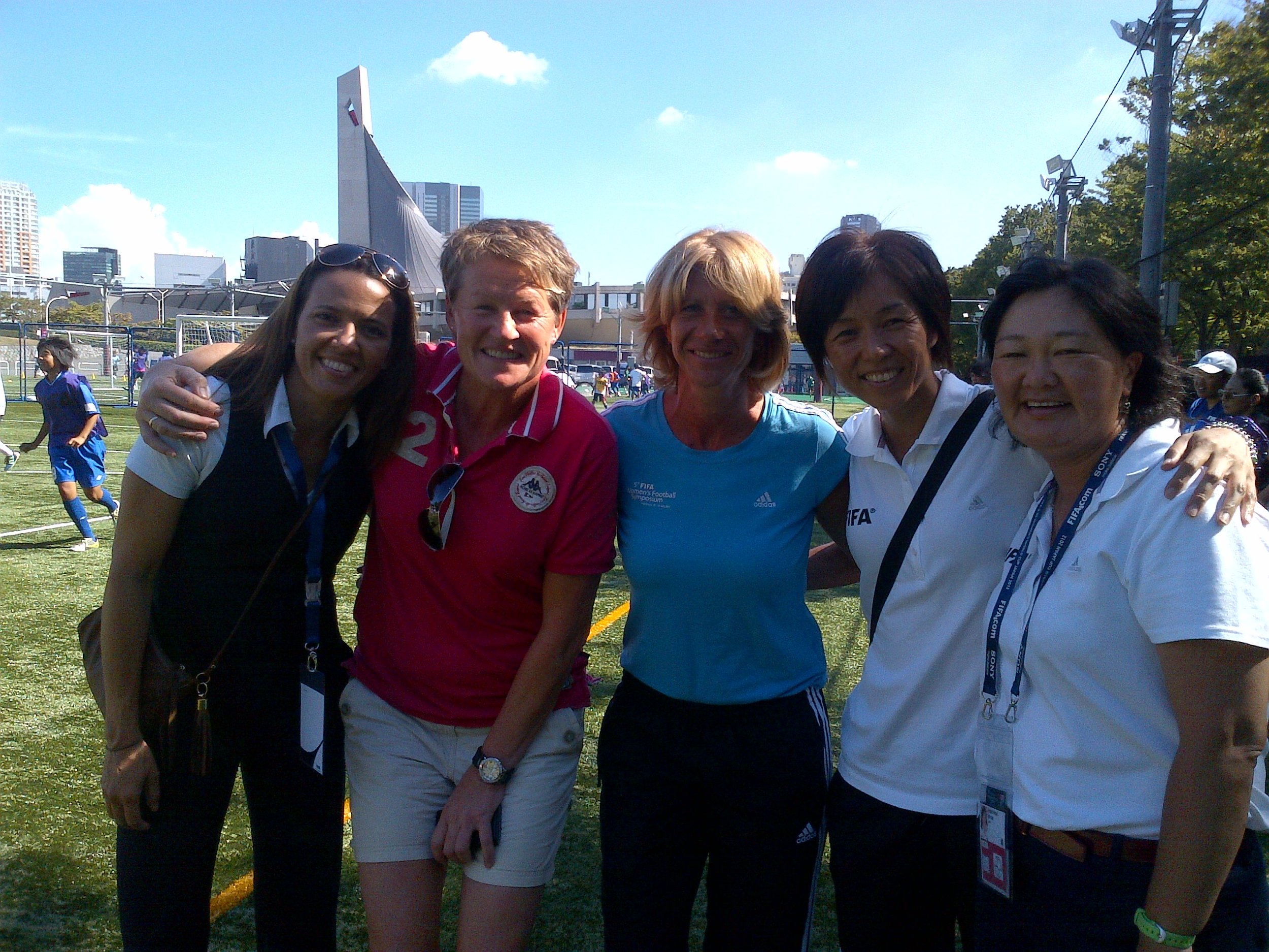 A group of five women smiling and standing close together outdoors at a sports event, with a city skyline and a stadium in the background.
