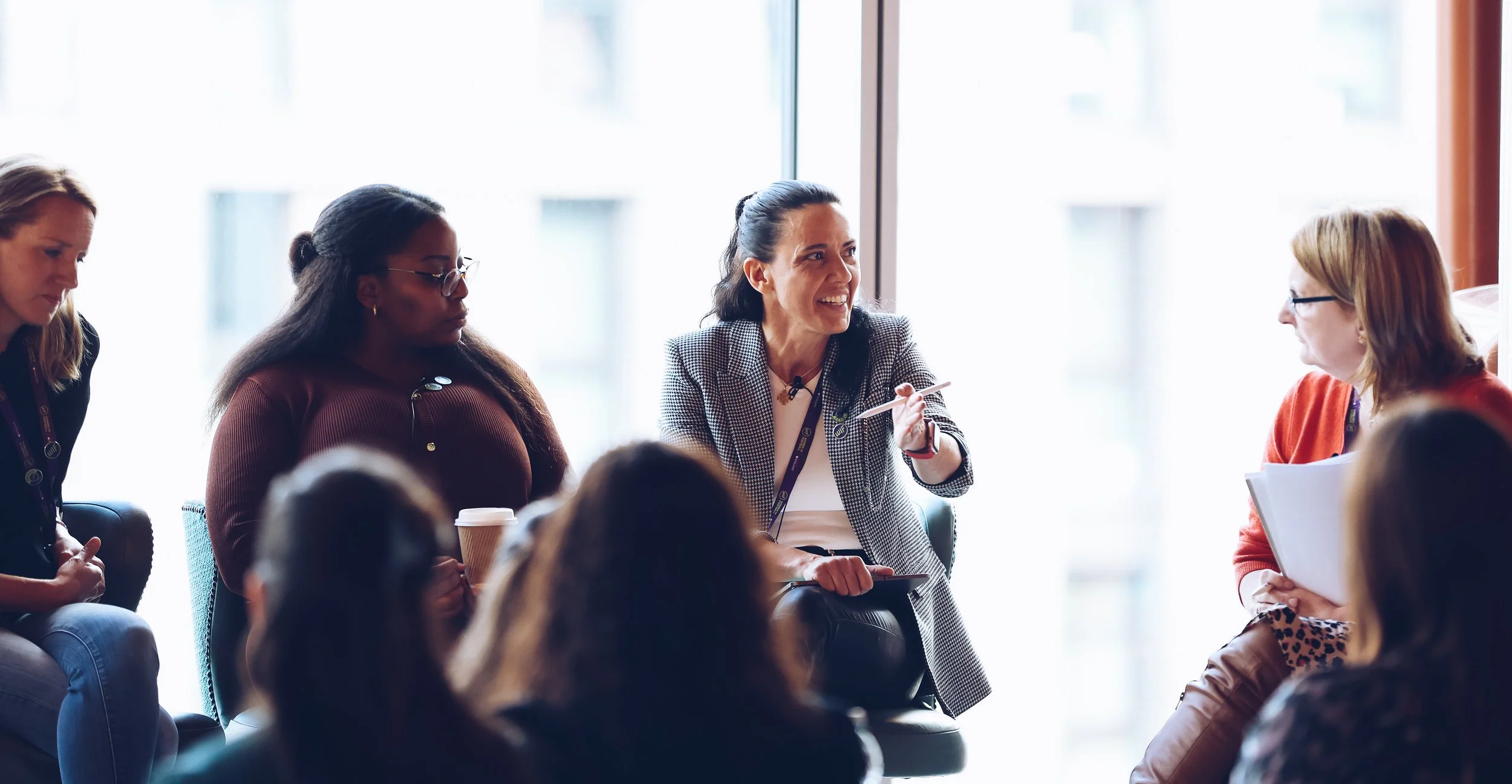 A group of women engaged in a discussion during a meeting or conference in a bright room with large windows.