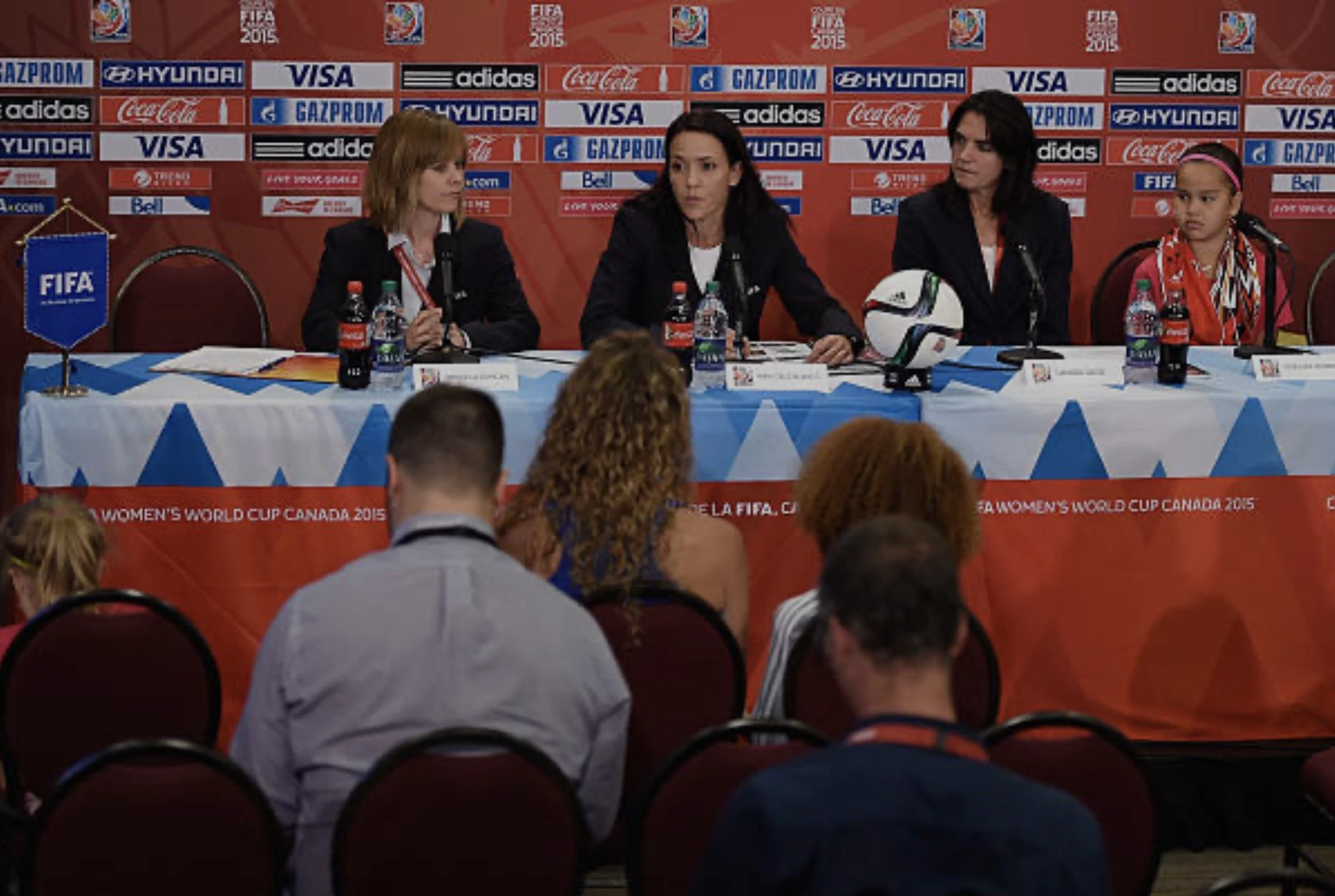 Two women speaking at a press conference with microphones, soda bottles, and a soccer ball on the table, and banners in the background.