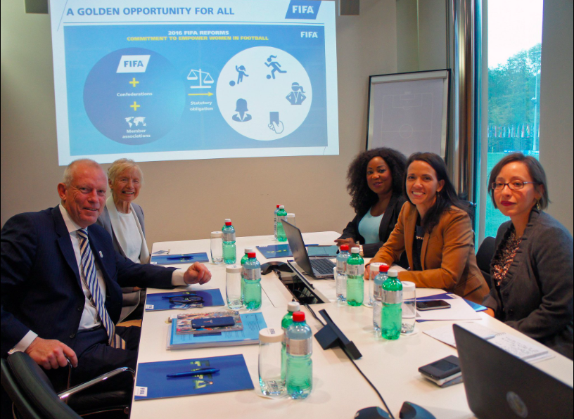 Five professionals sitting around a conference table in a meeting room, with a presentation slide explaining FIFA reforms projected on the wall behind them, and a large window showing an outdoor scene.