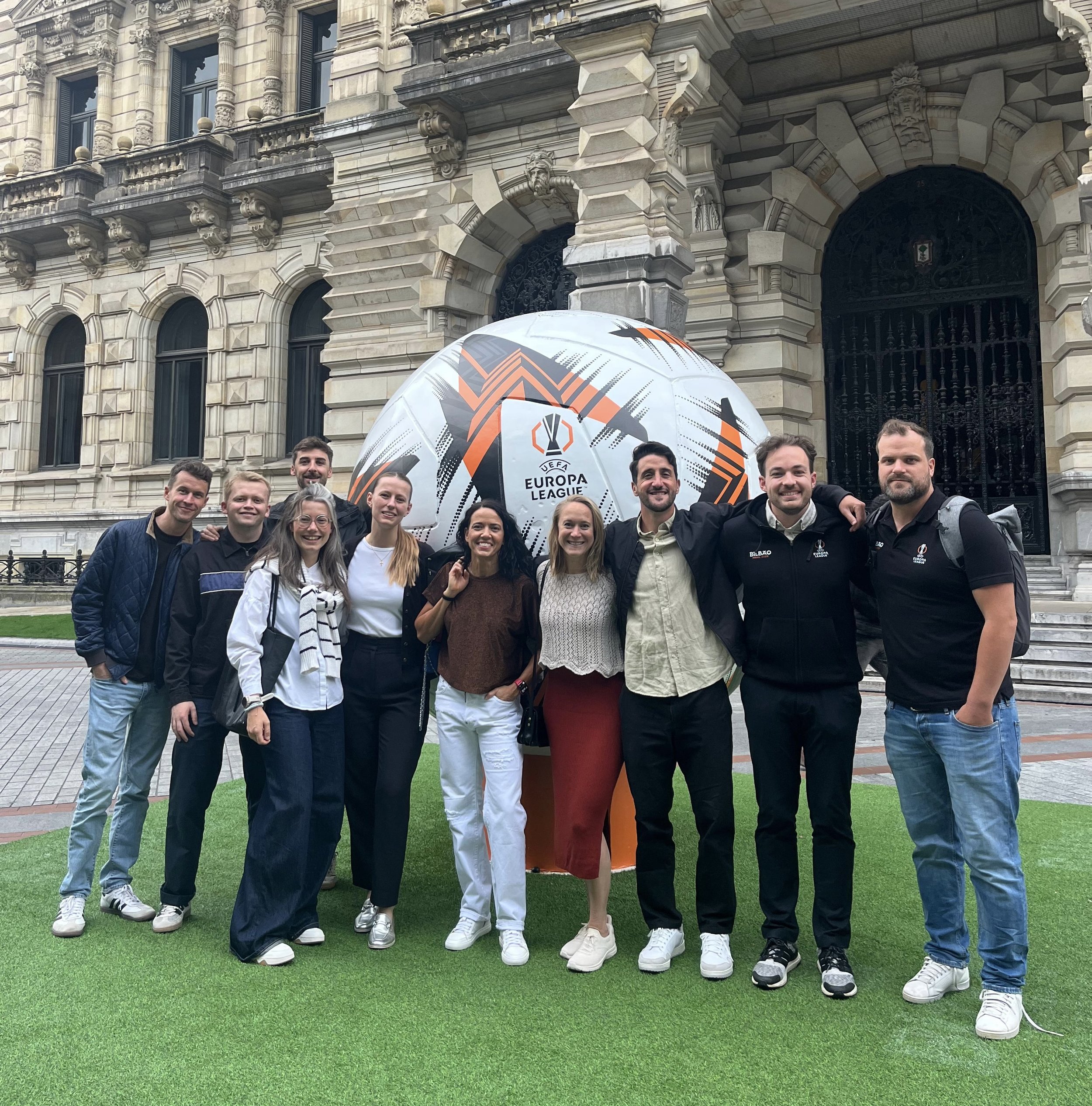 Group of people posing outdoors in front of an ornate historic building, holding a UEFA Europa League soccer ball.