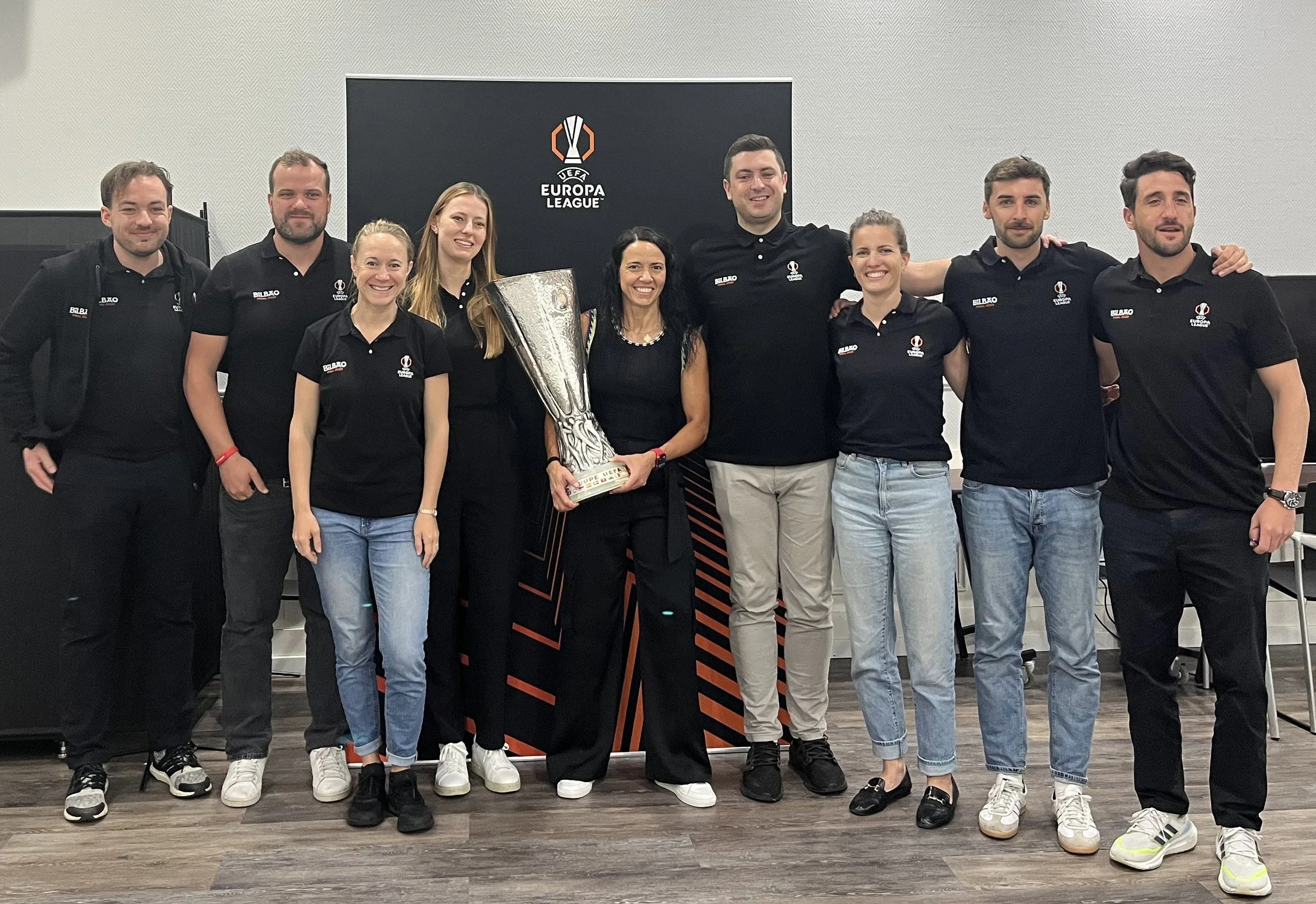 Group of nine people posing together indoors, some holding a UEFA Europa League trophy.