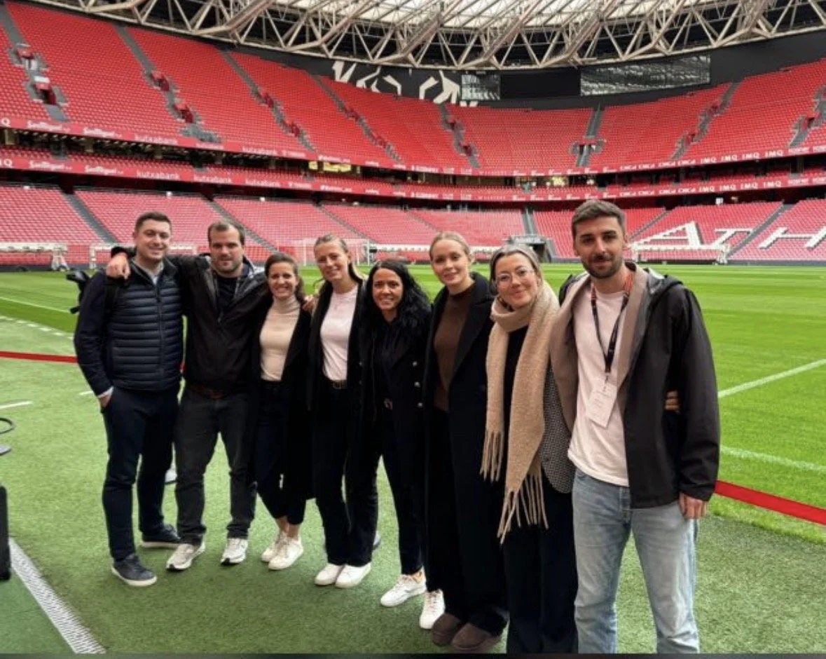 Group of eight people standing on a soccer field inside a stadium with red seats.