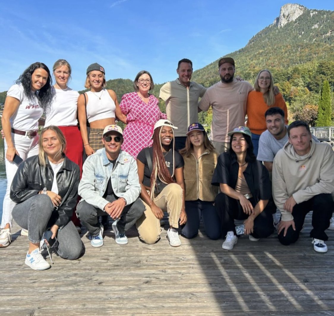 A group of fifteen people posing outdoors on a wooden deck with mountains in the background during daytime.