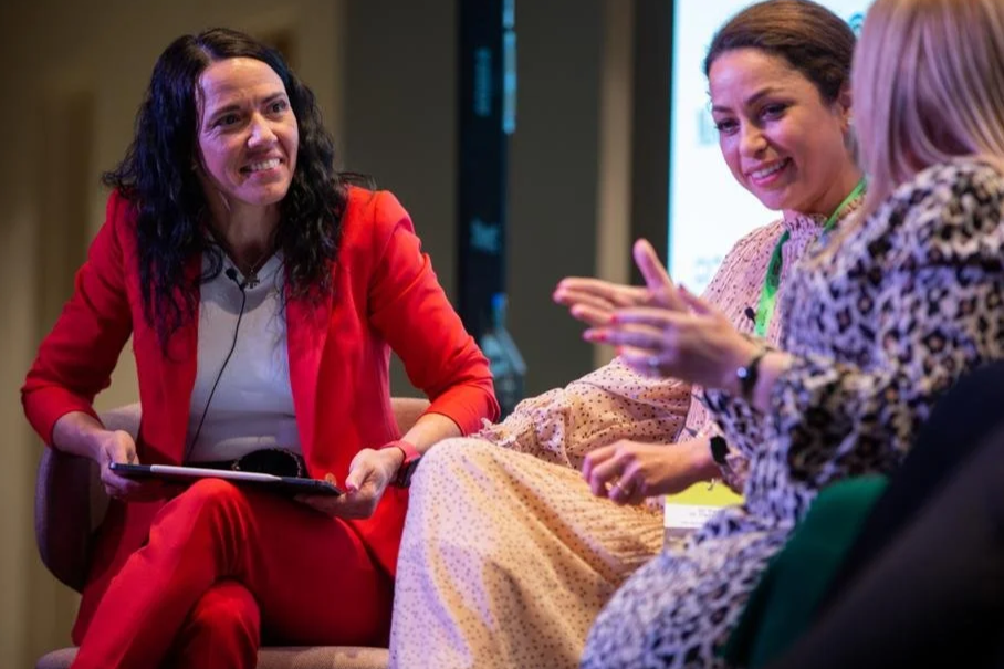 Three women sitting and engaged in a conversation at an indoor event, with one woman wearing a red blazer and holding a notepad, smiling and talking.