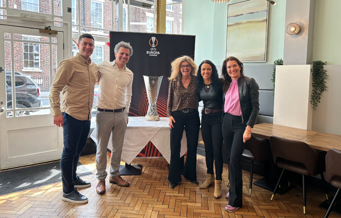 Group of five people standing indoors near a table with a trophy and a Europa League banner, smiling for the camera.