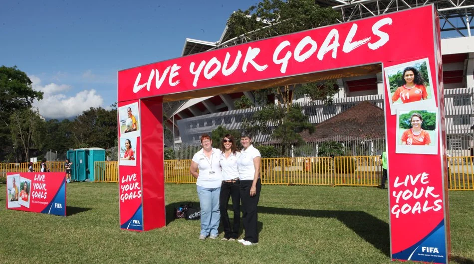 Three women standing under a large red archway with the words "LIVE YOUR GOALS" at an outdoor event. There are posters of female soccer players on the side of the arch and a smaller sign with the same message in the background. The event is organized by FIFA.
