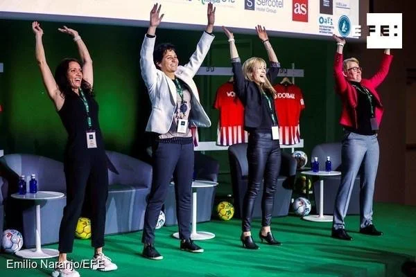 Four women standing on a stage at a conference or event, raising their arms and smiling. They are wearing formal business attire. Behind them are chairs, small tables with water bottles, and sports jerseys hanging on the wall.