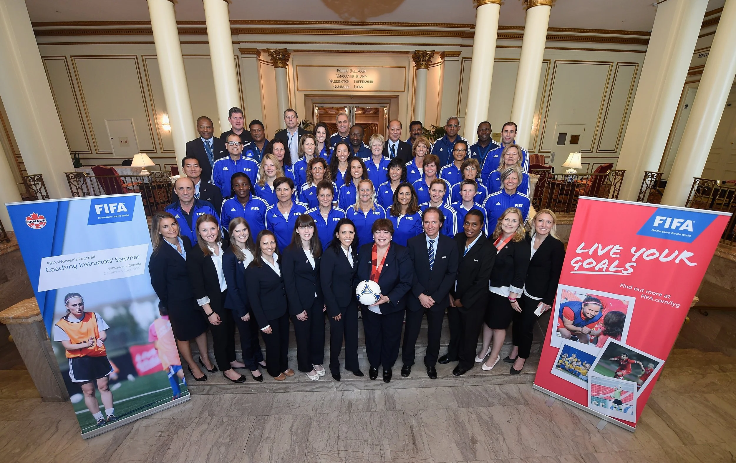 Group of people, mostly women and some men, posing for a photo at an indoor event. Several are wearing blue jackets with 'FIFA' logos, and some are in black suits. Two banners are displayed in front, one with a woman in a soccer uniform and the FIFA logo, and another with the slogan 'Live Your Goals' and images of women playing soccer.