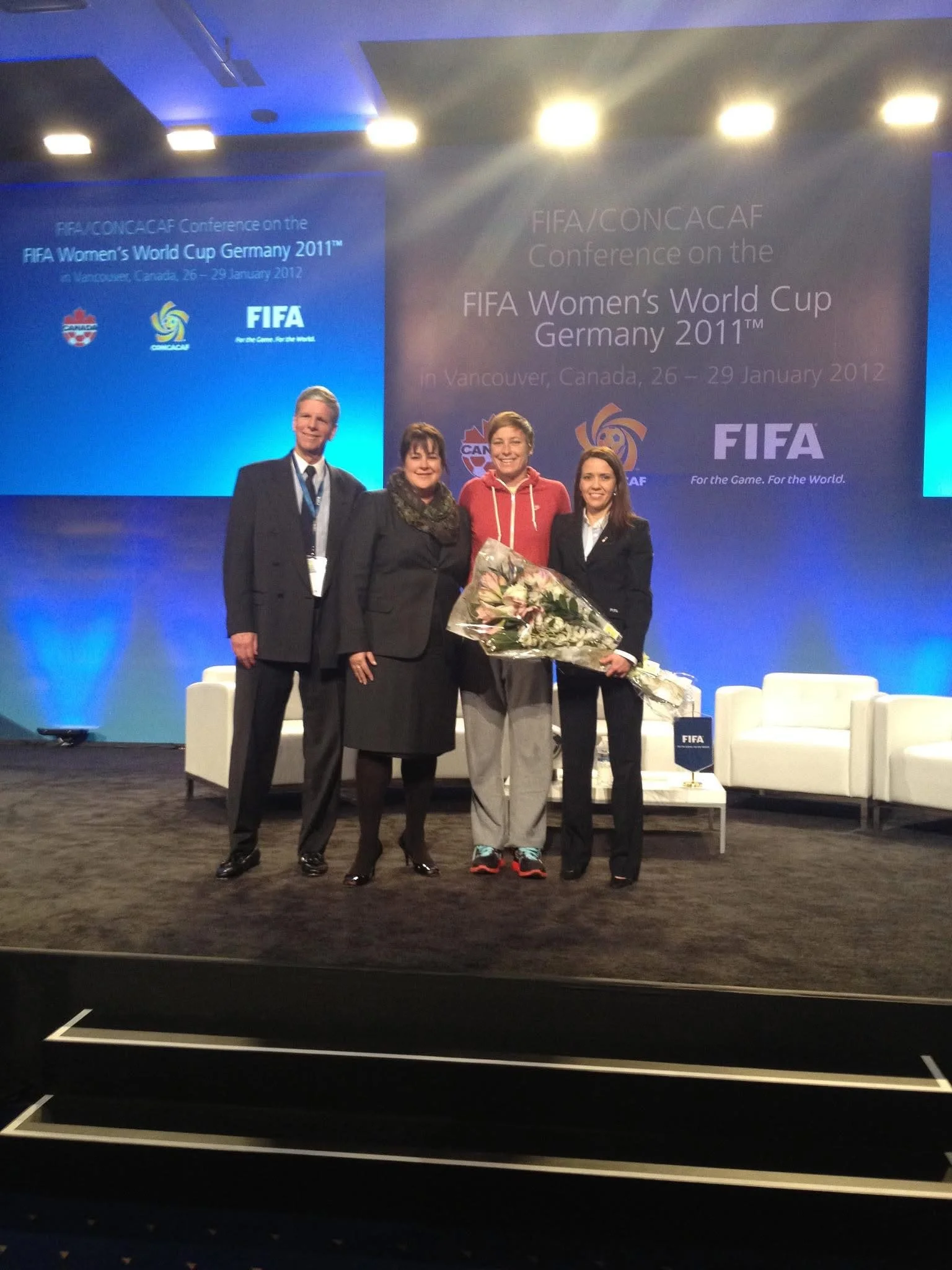 Four women standing on a stage, one holding a bouquet of flowers, at a conference about the FIFA Women's World Cup Germany 2011, with blue and black background screens displaying event information in Vancouver, Canada.