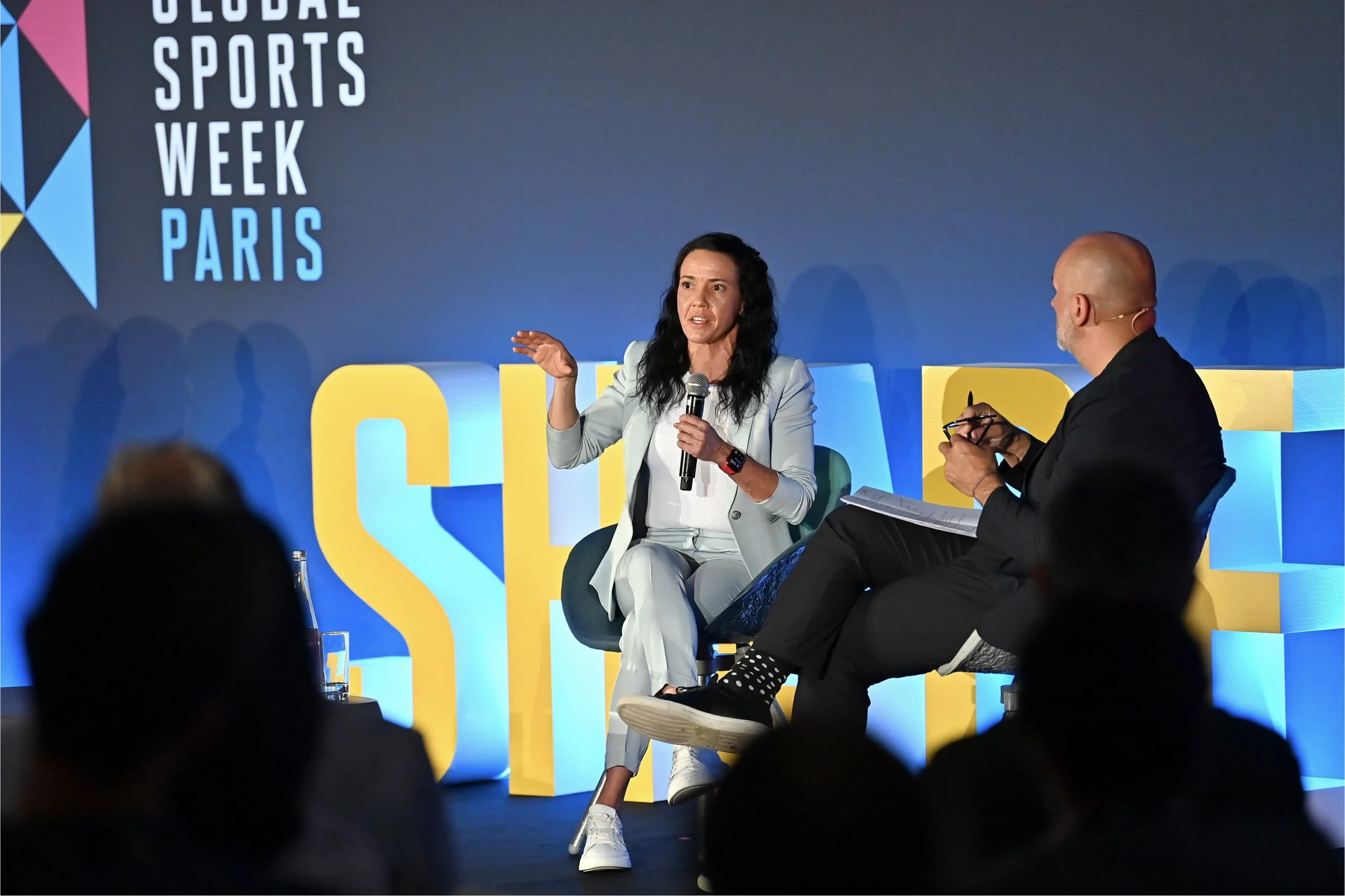 Two people are sitting on a stage with a blue background and the words 'Global Sports Week Paris' in large letters. The woman on the left is speaking into a microphone, gesturing with her right hand, and wearing a light blazer, white shirt, and white pants. The man on the right, bald with a beard, is wearing a black suit, holding a pen and a notepad, and looking at the woman. There are some audience members' silhouettes in the foreground.