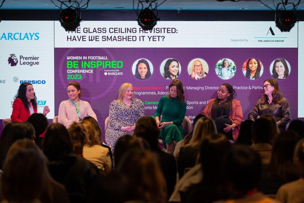 Panel of six women sitting on stage during a conference, with an audience in front. Behind them is a large screen displaying event information and speaker photos.