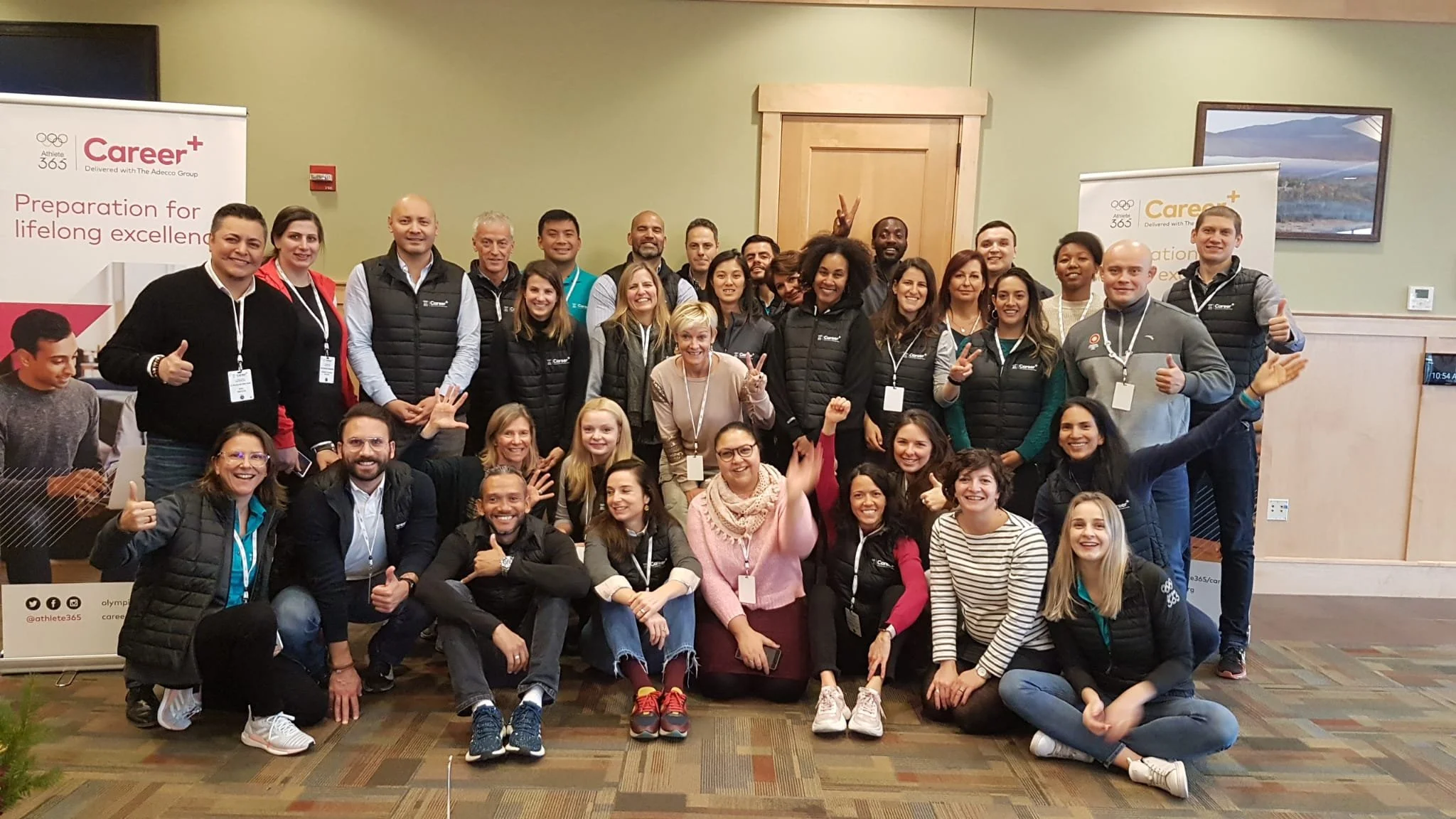 Group of diverse people posing together indoors for a photo at a professional event, some making peace signs or thumbs up, with banners for Career+ and Athlete 365 in the background.
