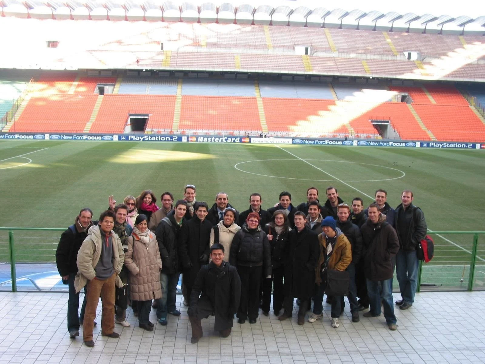 Group of people standing on a balcony overlooking a soccer field inside a stadium, with empty red and blue seats and advertisement banners.