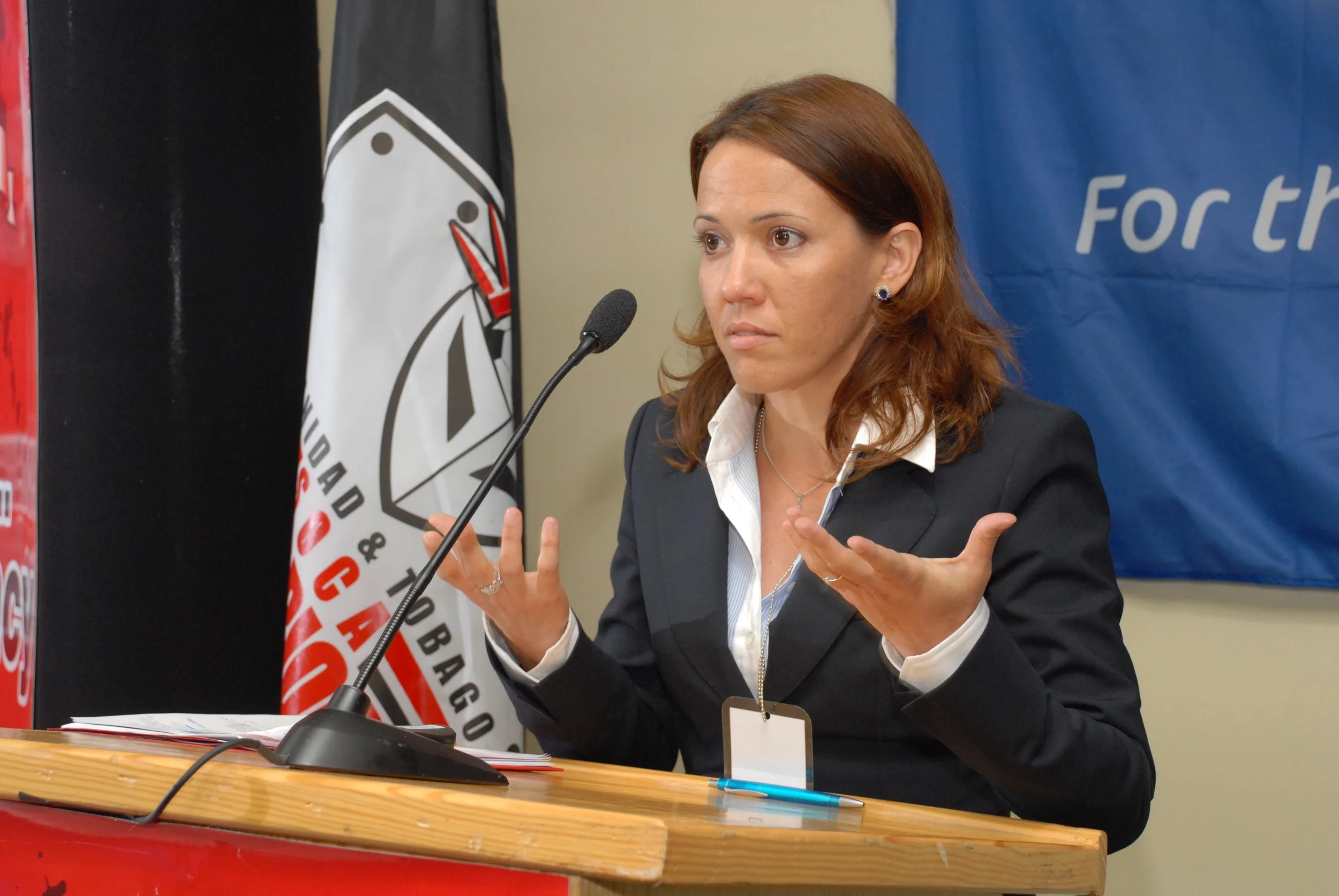 A woman with reddish brown hair speaking at a podium, gesturing with her hands, dressed in a business suit and white shirt, with flags and banners in the background.