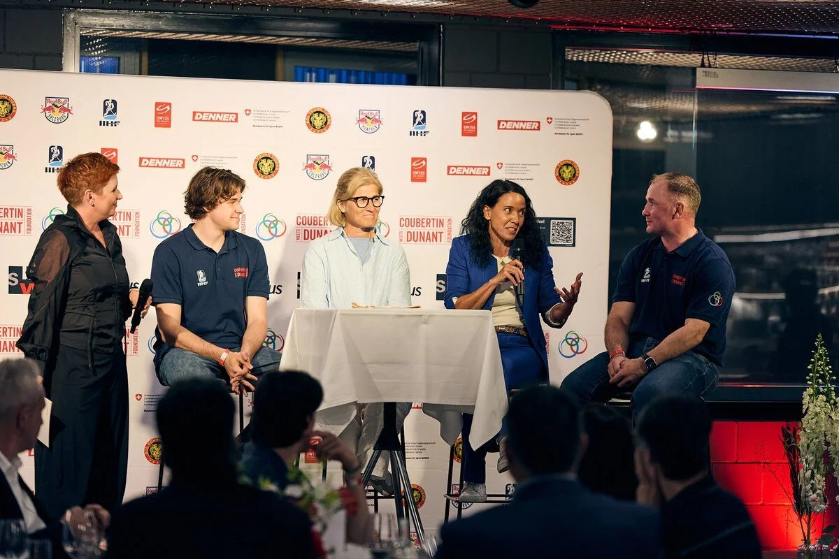 Panel discussion with five people on stage, four seated and one standing, at an event with a backdrop featuring logos such as Red Bull, Denner, and others.