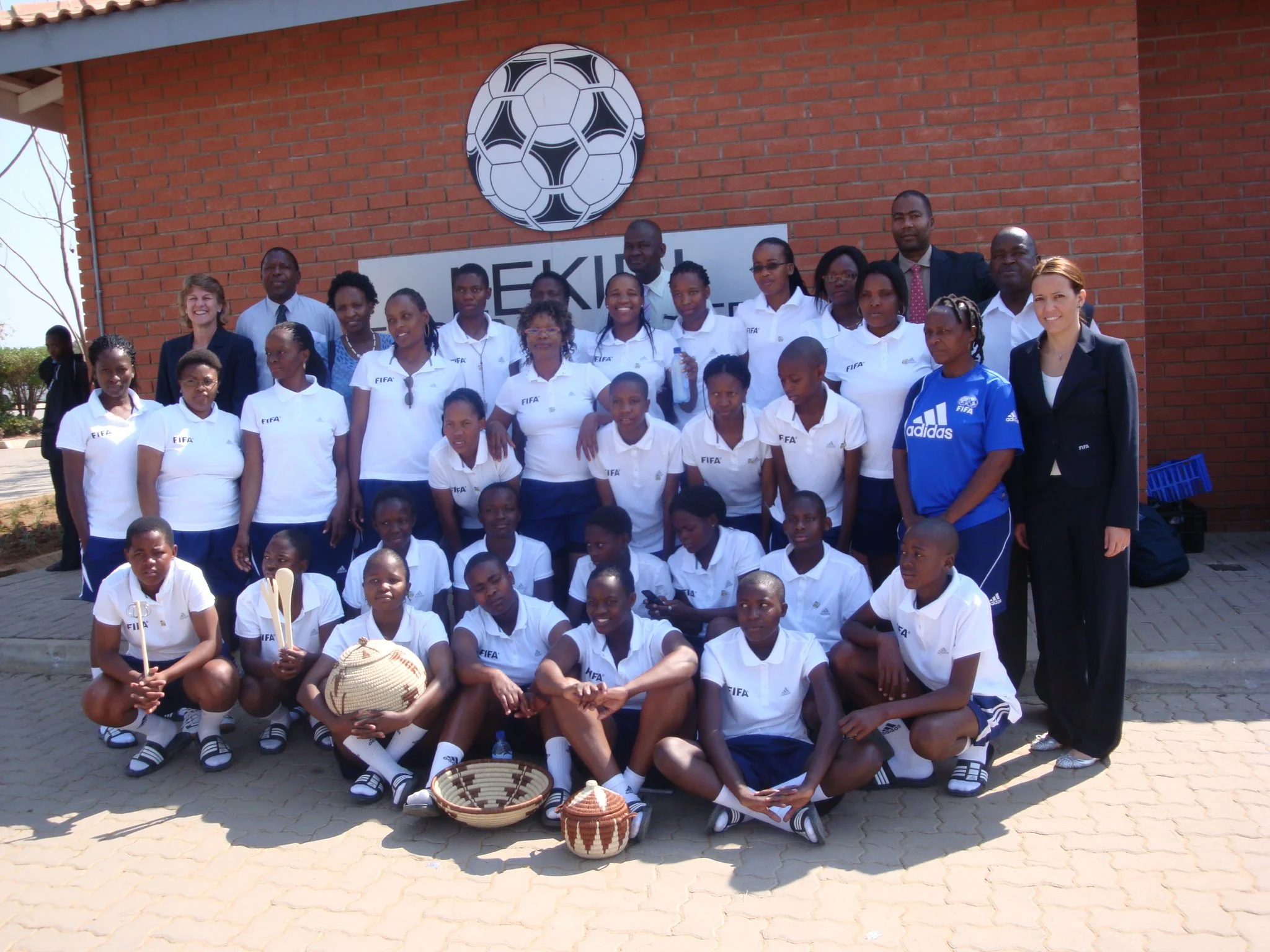 Group of children and adults posing in front of a brick wall with a soccer ball logo, some wearing white FIFA shirts, a few holding baskets, on a paved outdoor area.