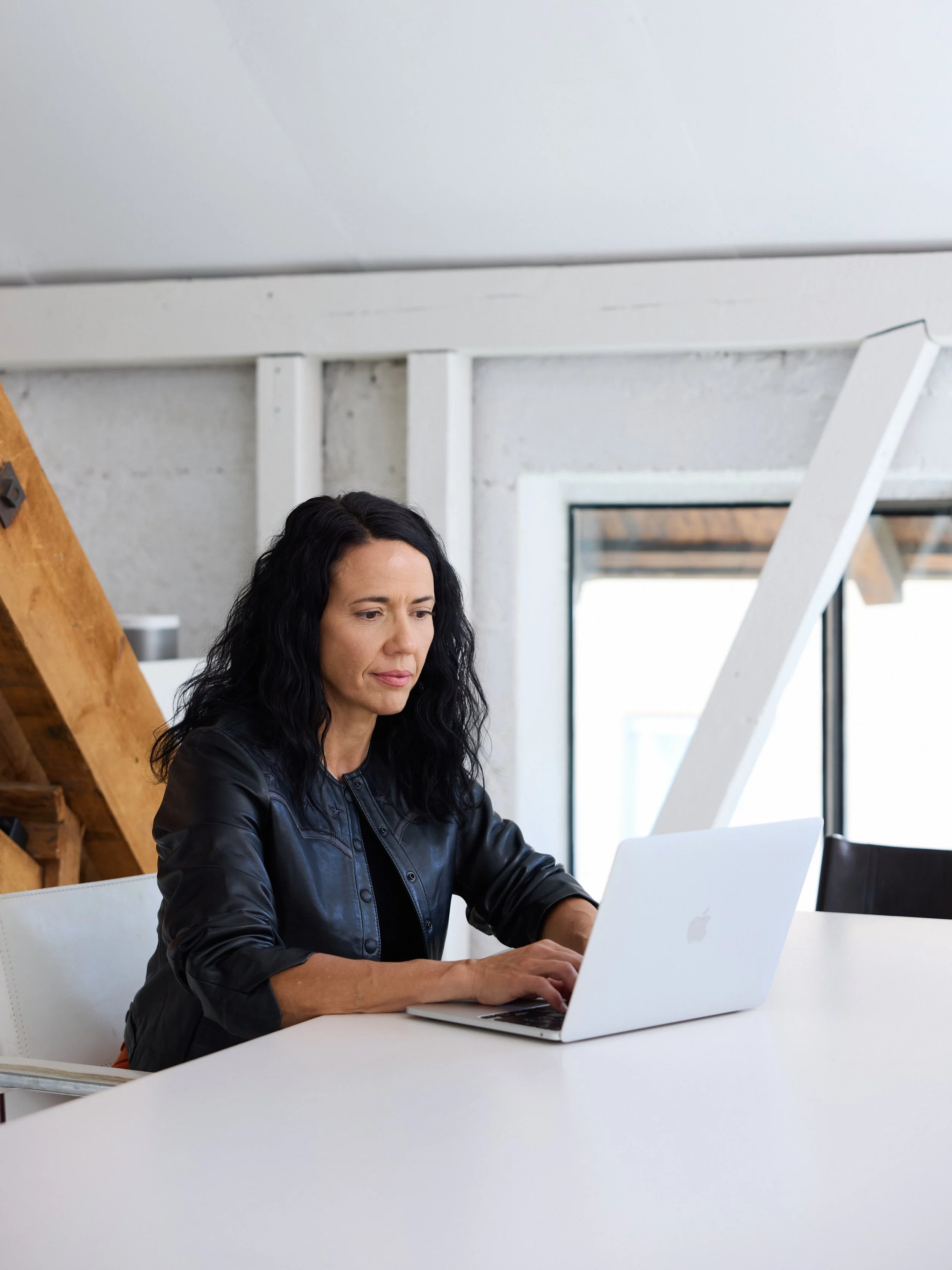 A woman with black wavy hair, wearing a black leather jacket, working on a silver laptop at a white table in a modern room with white walls, a window, and wooden beams.