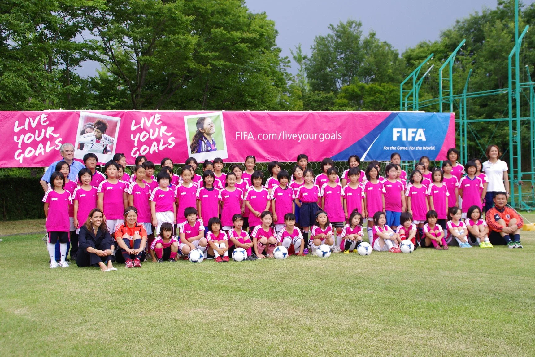 A large group of young girls in pink sports uniforms, along with their coaches and adult supervisors, posed for a team photo on a soccer field during daytime. Behind them is a pink banner with photos of female soccer players and the text 'LIVE YOUR GOALS' and 'FIFA.com/liveyourgoals.' The background features green trees and overcast sky.