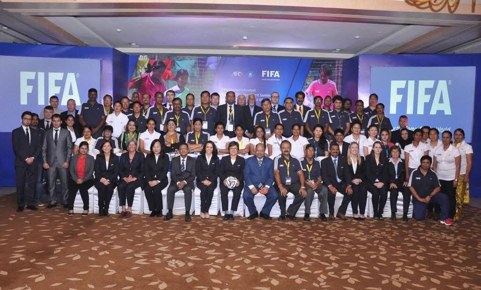 Group photo of diverse FIFA officials, delegates, and staff in a conference room with FIFA branding on large screens in the background.