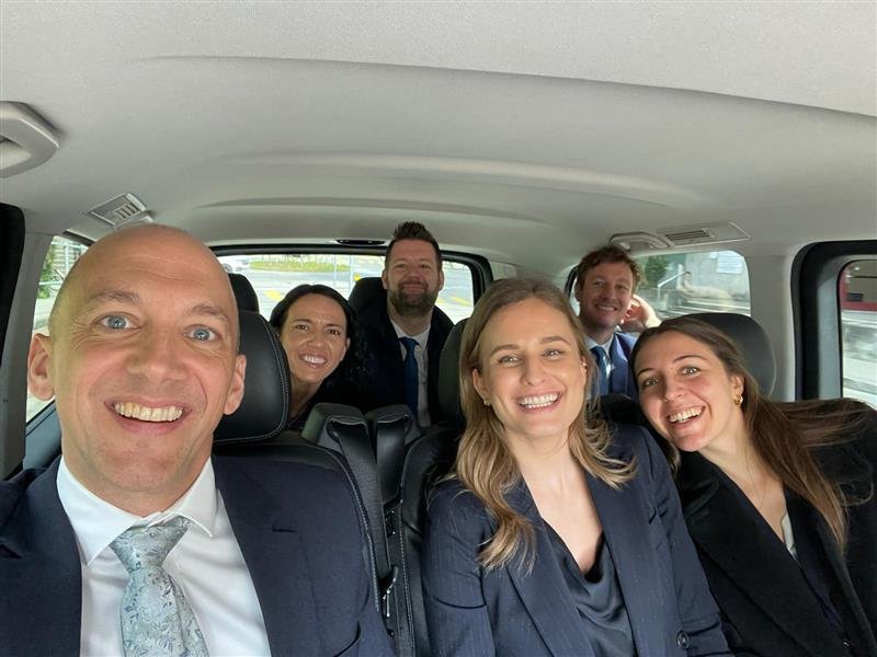 Six professionally dressed people smiling inside a vehicle, taking a group selfie.