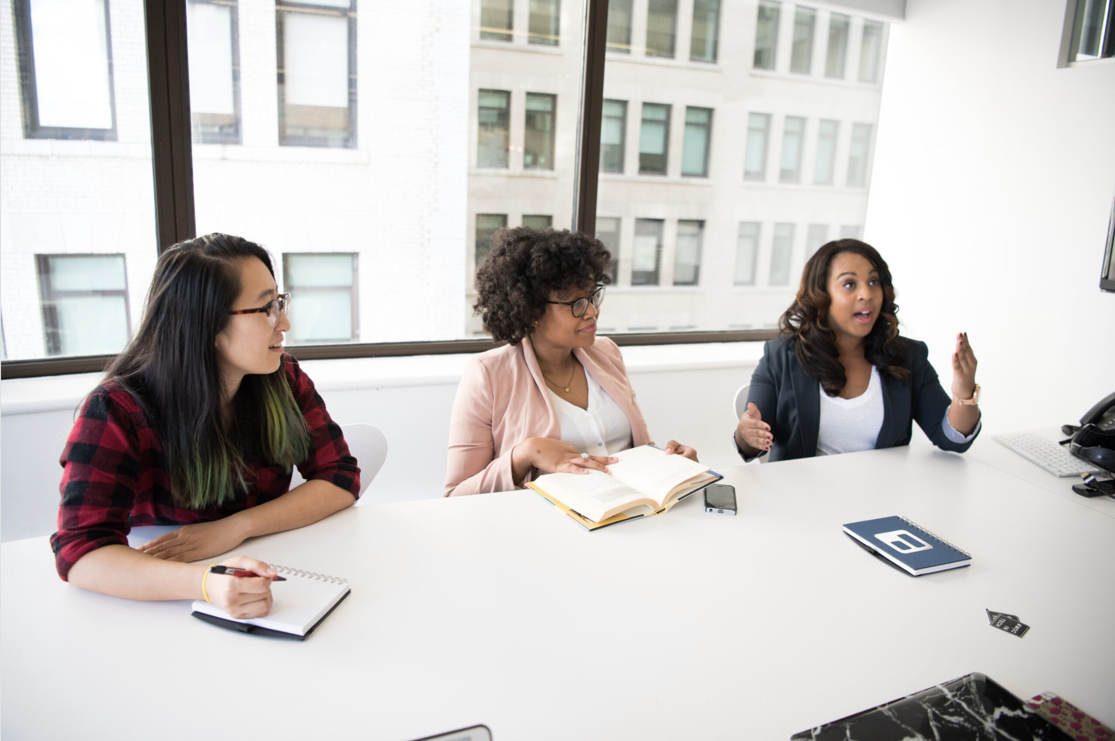 Three women sitting at a white table in a modern office with large windows, engaging in a discussion. One woman is speaking and gesturing with her hand, while others are listening and taking notes.