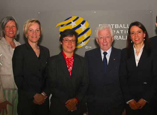 Five women posing in front of a sign that reads 'Football Federation Australia,' with a blue and yellow logo.