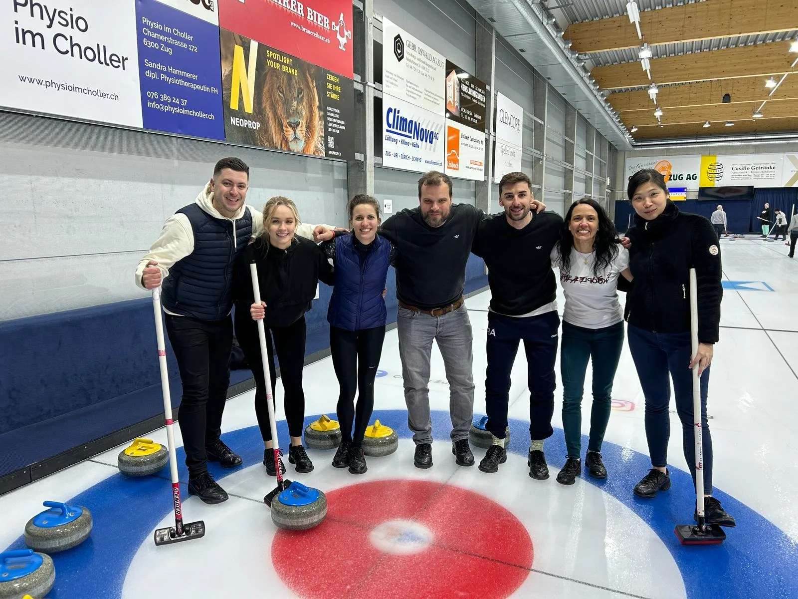 Group of seven people standing on an indoor ice curling rink, smiling, with curling stones in front of them, and banners on the wall behind.