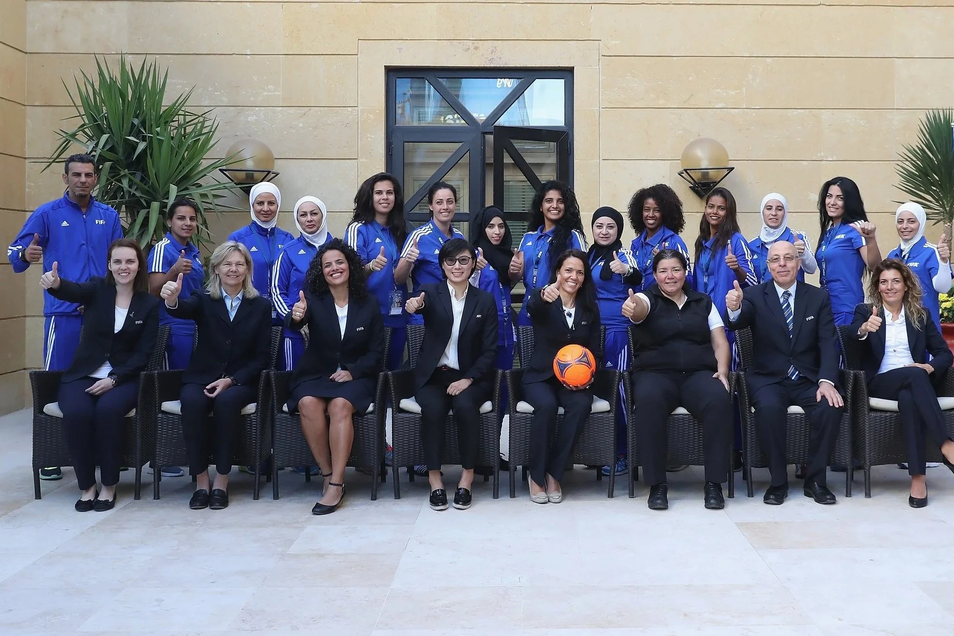 Group of women and men, some in sportswear and some in formal business attire, posing outdoors in front of a beige wall with large plants on each side, giving a thumbs-up gesture.