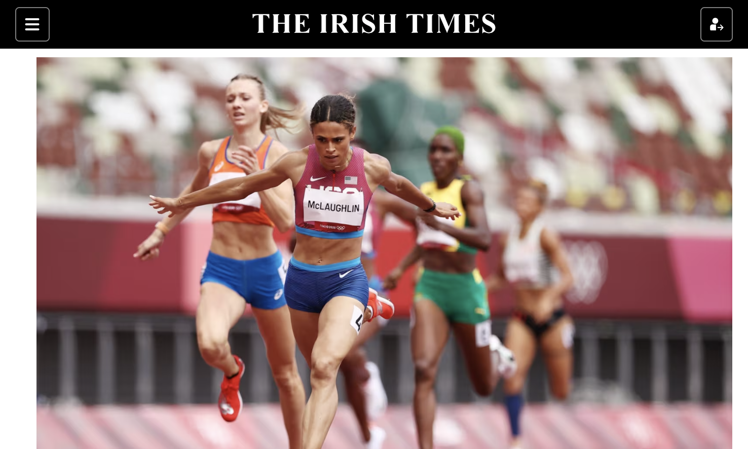 Female athletes competing in a track race, with one runner in the foreground wearing a pink top and blue shorts, and others behind her, on an outdoor stadium track.