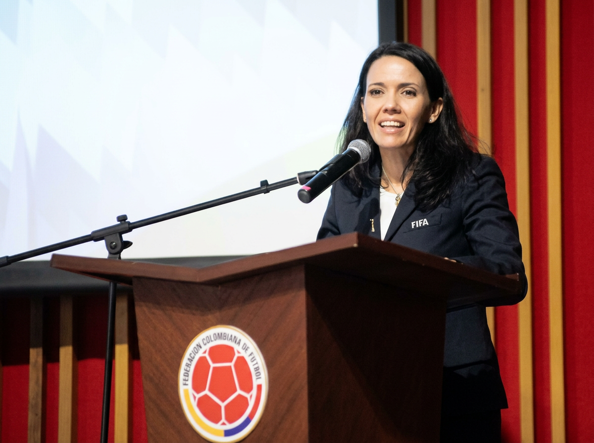 Woman speaking at a podium with a microphone, wearing a black suit with 'FIFA' on the chest, in front of a screen and red wall.