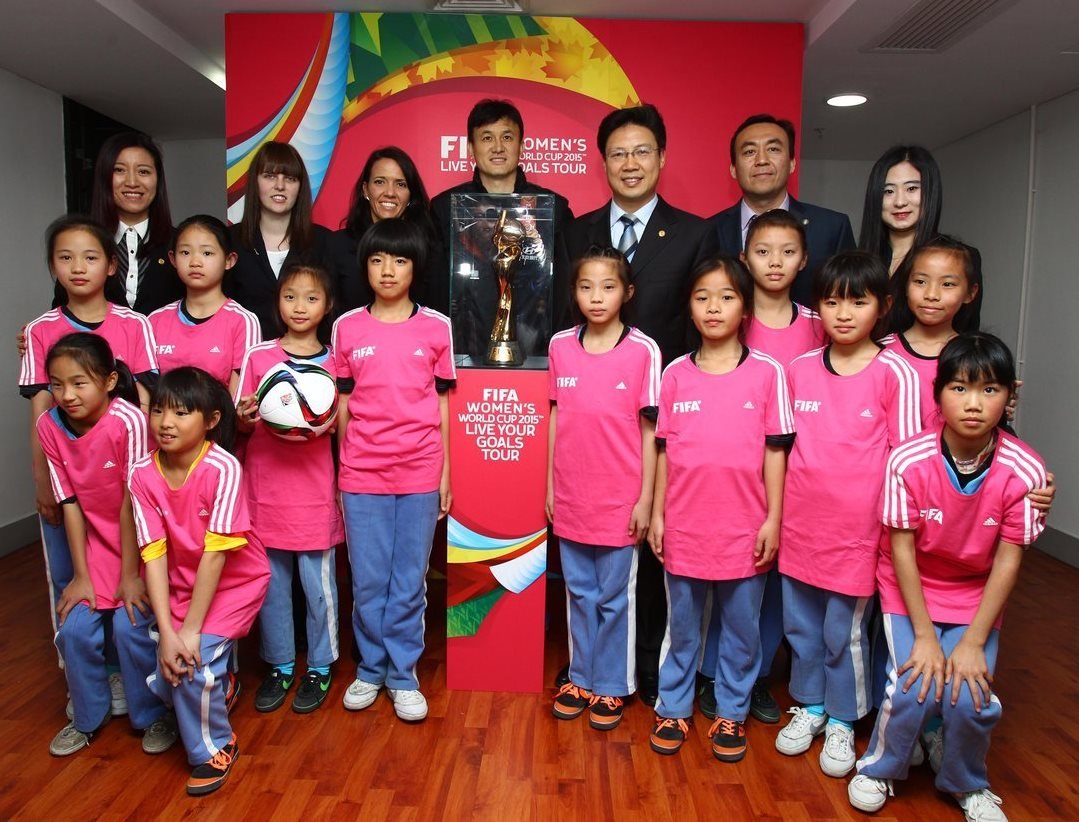 Group photo of young girls in pink soccer uniforms with FIFA logos, posing with officials and a FIFA Women's World Cup trophy at the 2015 tour event.
