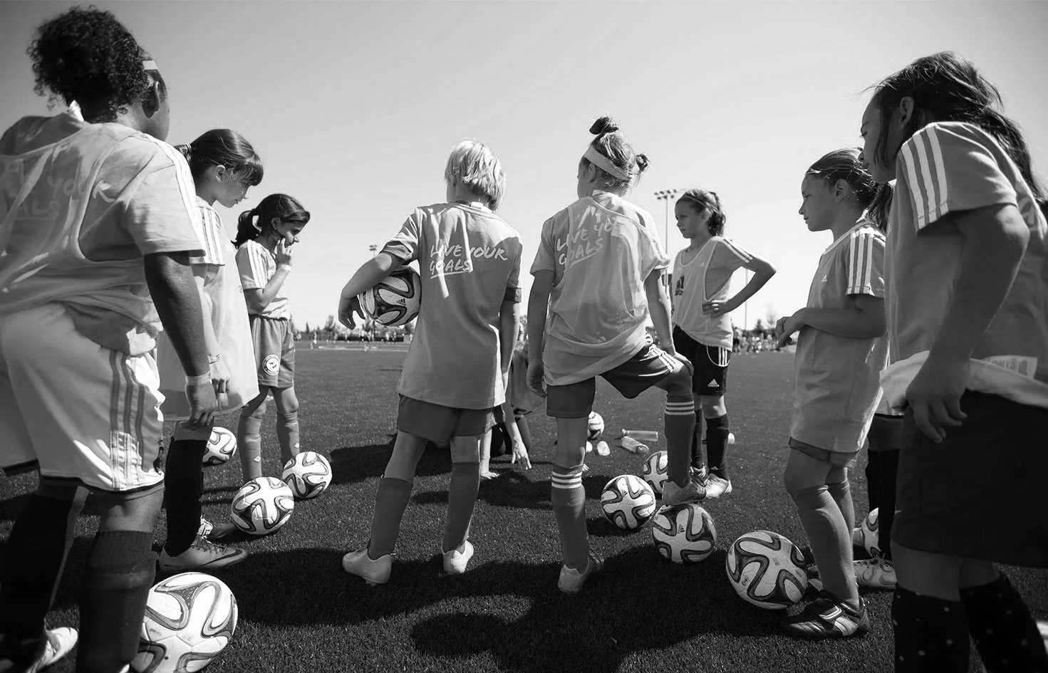Group of young girls in soccer uniforms standing in a circle on the field, with soccer balls at their feet, during practice or team meeting.