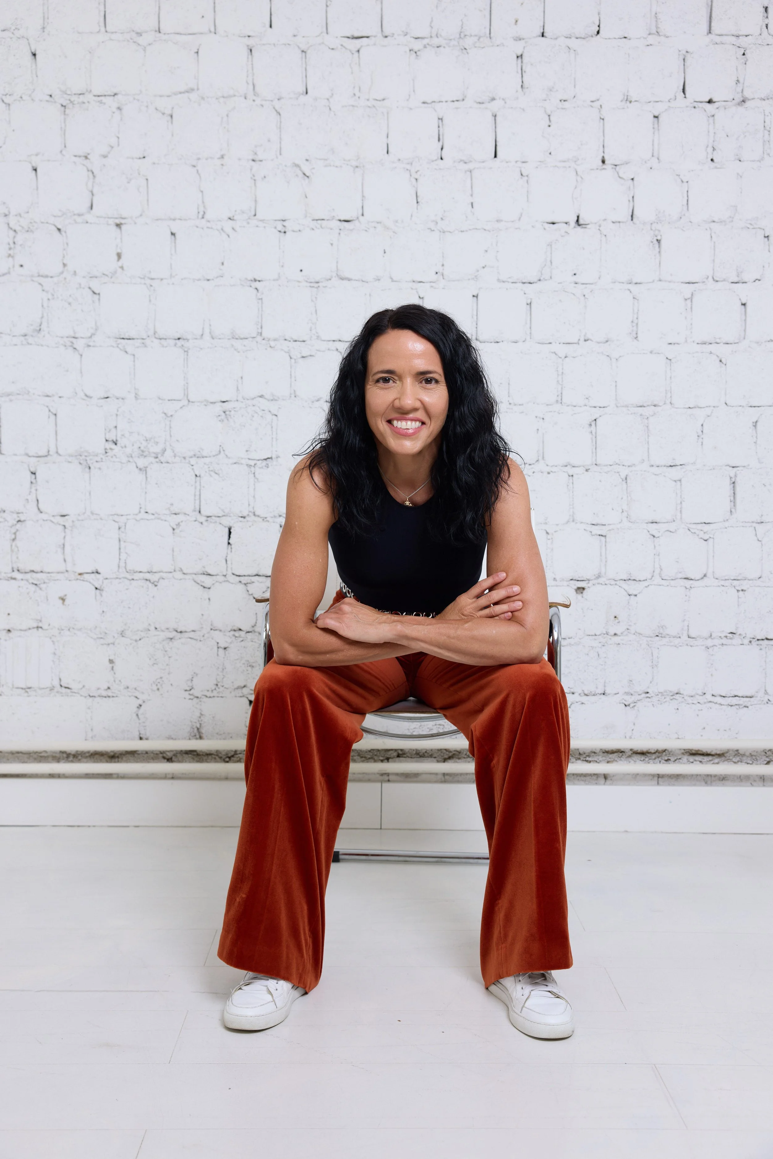 A woman with long curly black hair is sitting on a chair against a white brick wall, smiling at the camera, wearing a black sleeveless top, rust-colored wide-leg pants, and white sneakers.