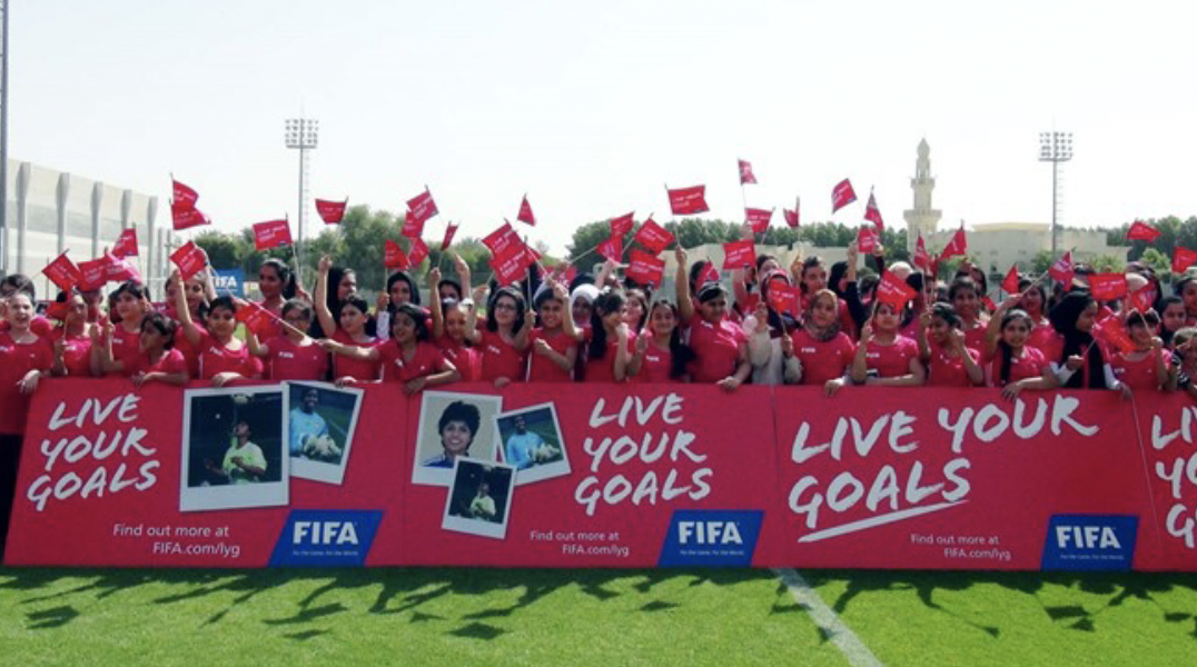 Group of women and girls holding red flags and banners at a FIFA event on a soccer field with a large FIFA sign and a lighthouse in the background.