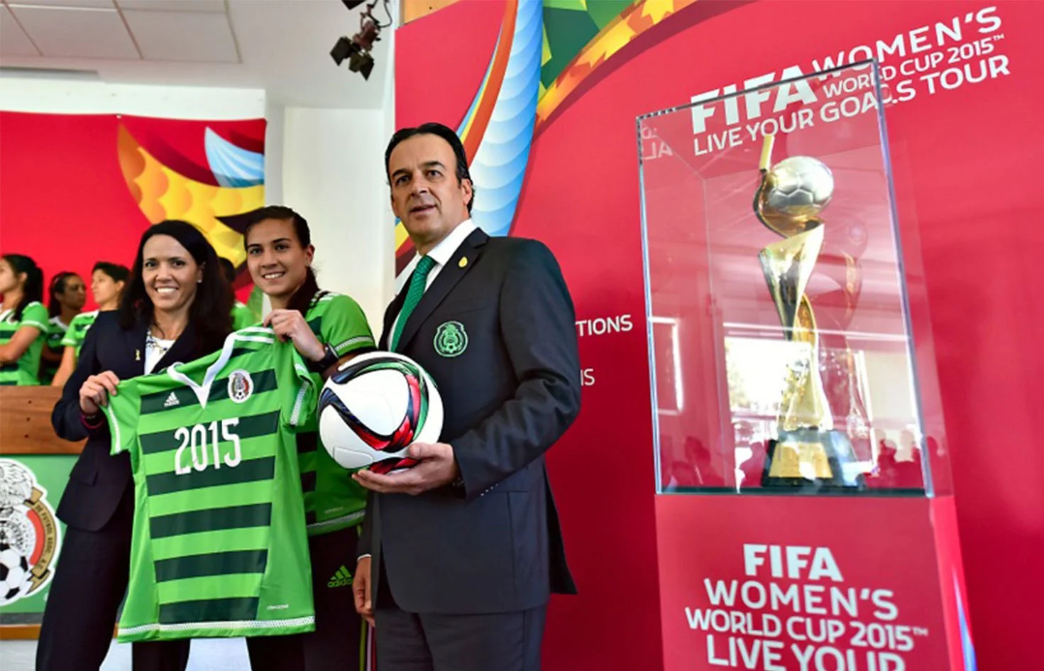 Three people standing in front of a red background with FIFA Women's World Cup 2015 branding. The woman on the left is holding a green Mexico national soccer team jersey with the year 2015 on it. The man on the right is holding a white and black soccer ball.