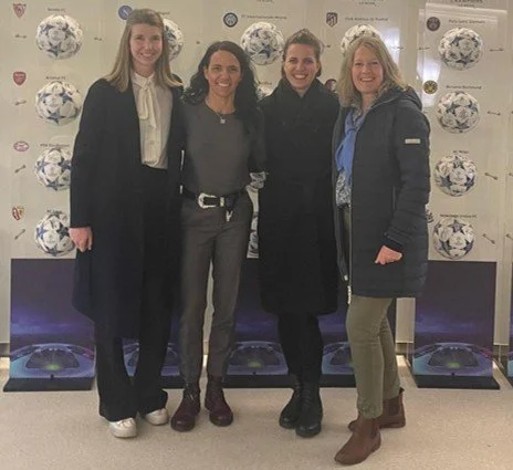Four women standing together indoors, smiling for a photo, with a backdrop featuring soccer balls and logos.