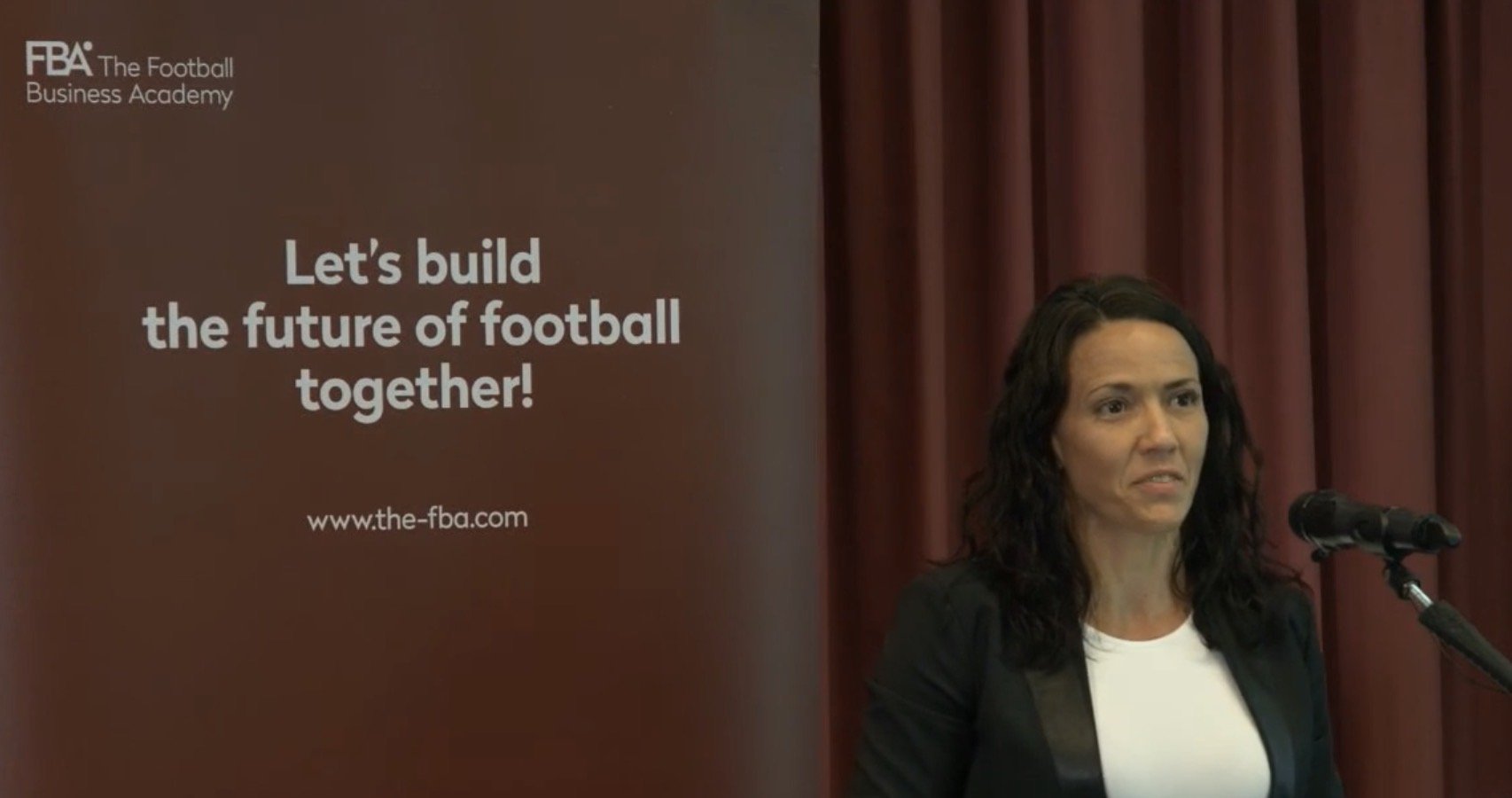 Mayi speaking at a podium with a brown curtain backdrop, next to a large brown banner that reads "Let's build the future of football together!" and includes the website www.the-fba.com. The banner has the logo for the Football Business Academy.